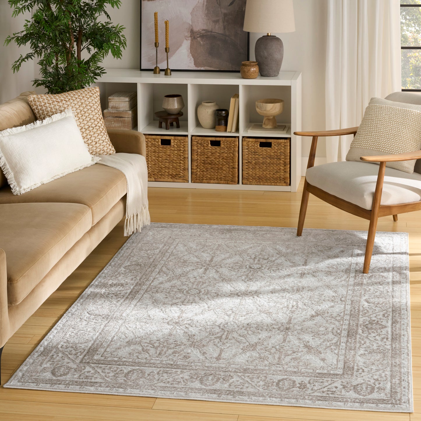 Neutral living room with beige sofa, light wood floors, gray patterned rug, white cubby shelf with wicker baskets, chair.