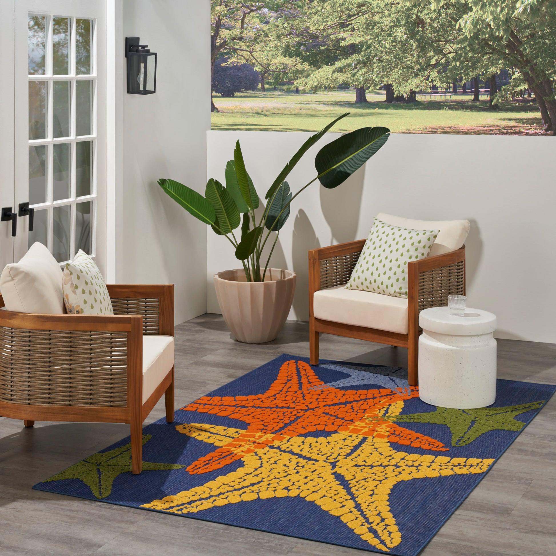Outdoor patio with two brown wicker armchairs, white cushions, vibrant starfish rug, large potted plant, white side table.