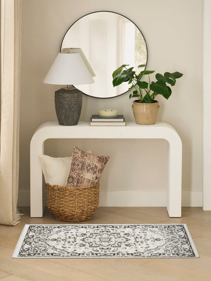 Round mirror over a white curved console with a dark textured lamp, potted plant, books; wicker basket with cushions; rug.