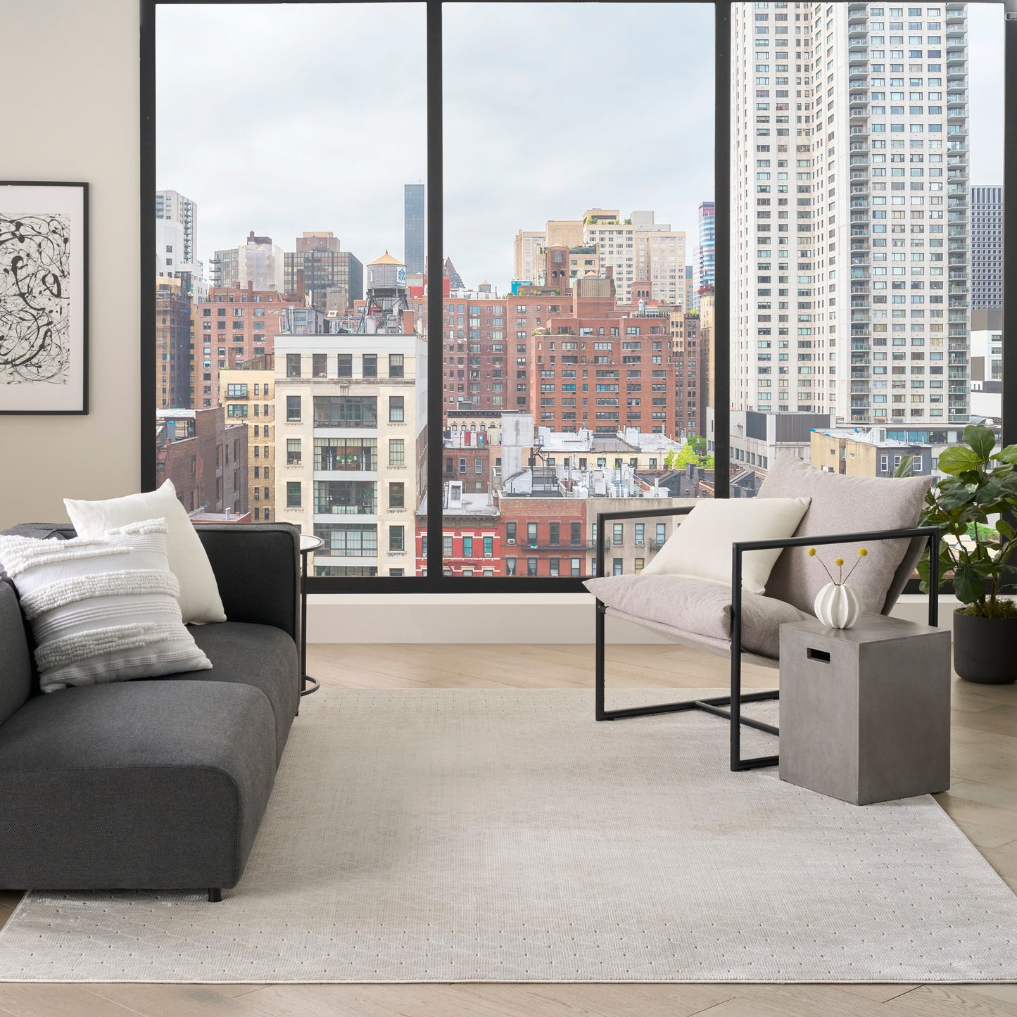 Modern living room with city-view windows, dark gray sectional, beige rug, metal-framed lounge chair, concrete side table.
