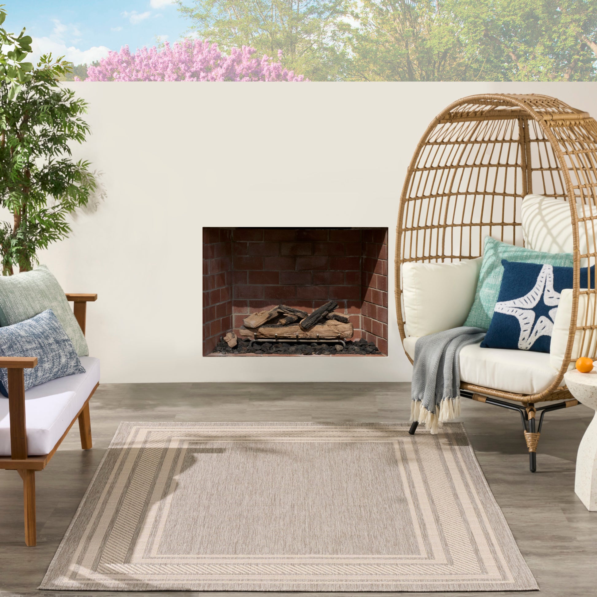 Outdoor patio with beige woven rug, brick fireplace, rattan egg chair with cushions, wooden lounge chair & potted plant.