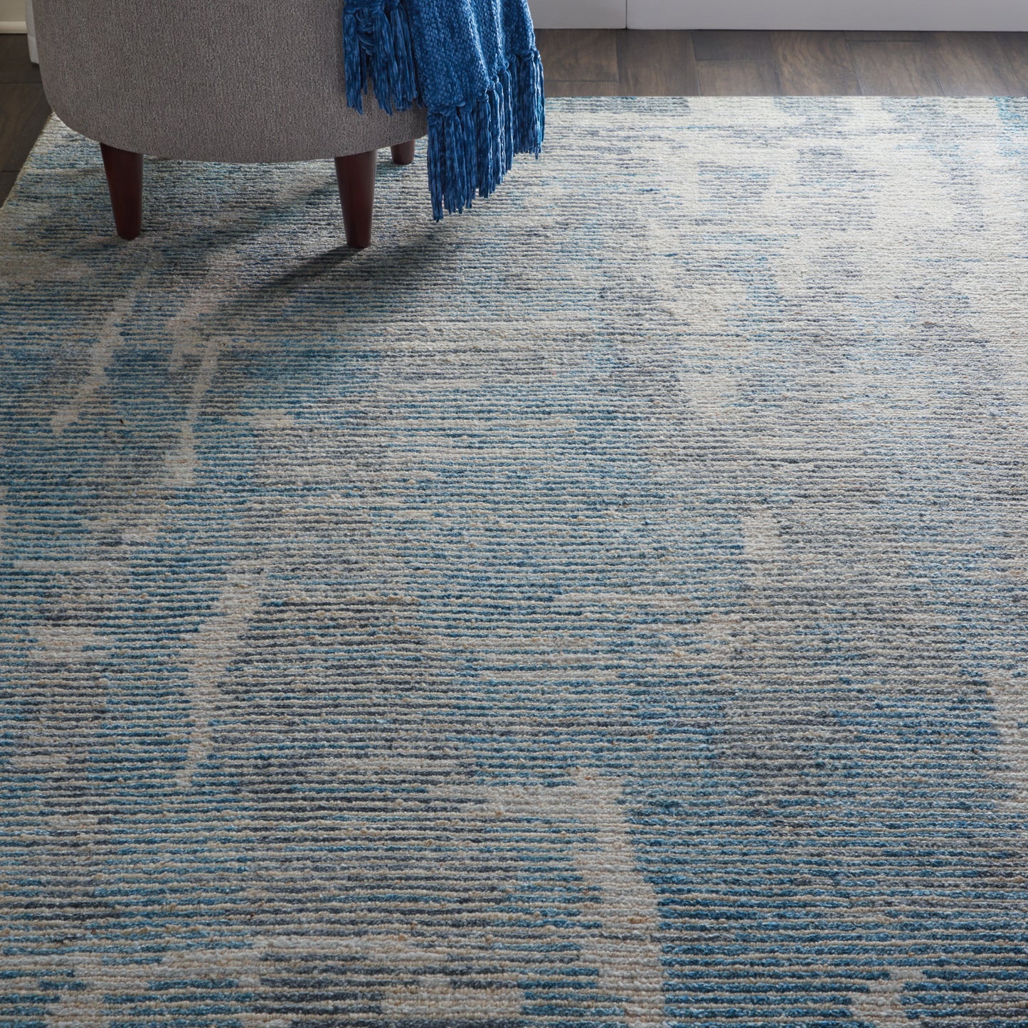 Blue-gray textured area rug with abstract wavy pattern beside a gray chair and a blue fringe throw.