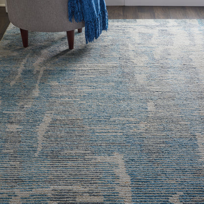 Blue-gray textured area rug with abstract wavy pattern beside a gray chair and a blue fringe throw.