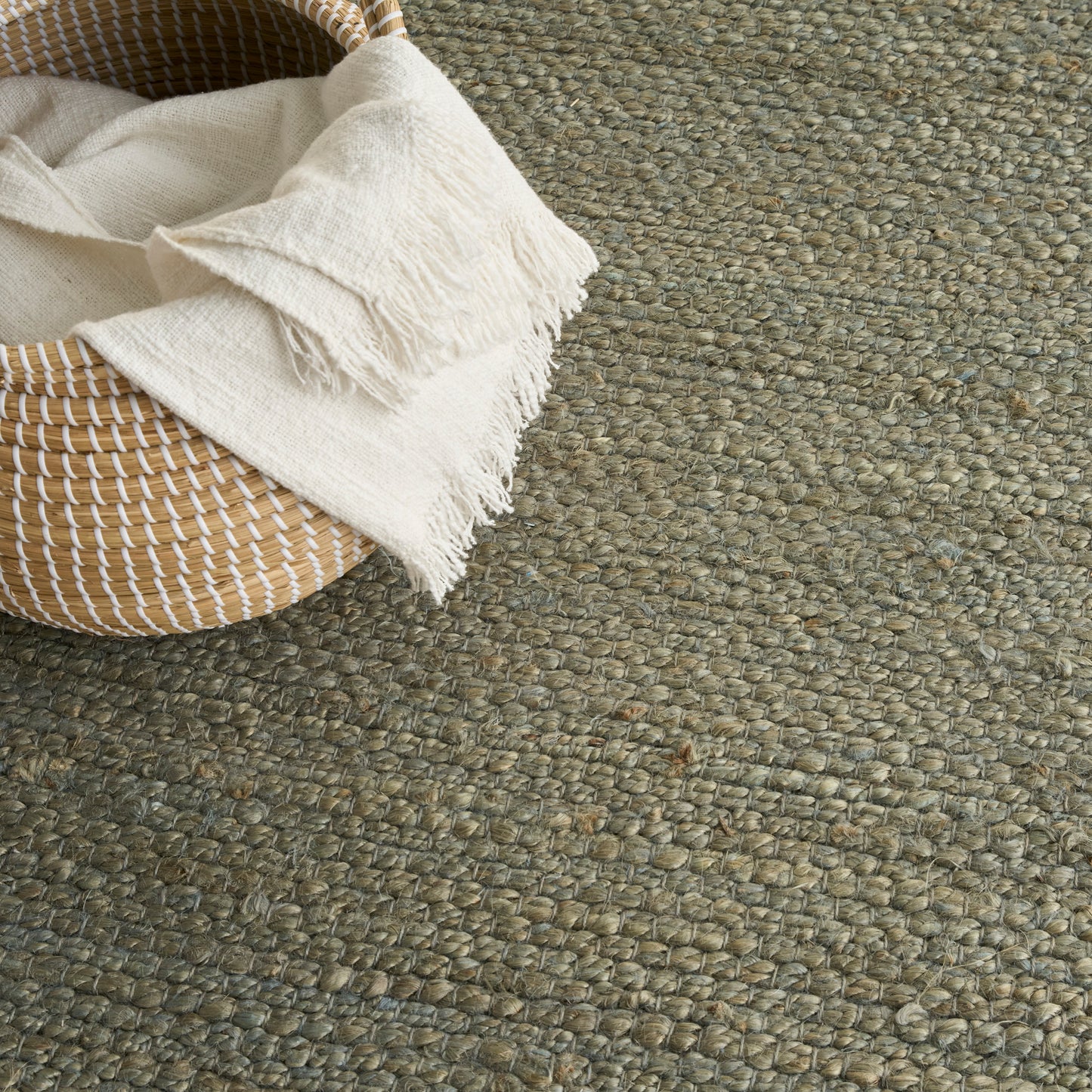 Round wicker basket with ivory fringed blanket resting on a chunky textured beige rug.