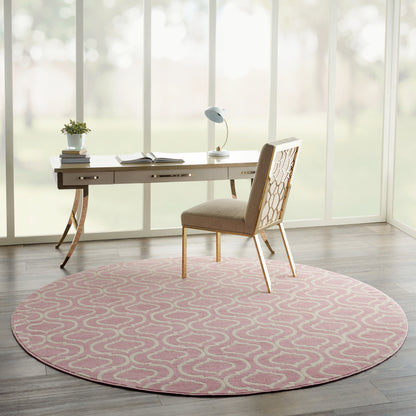 Round pink rug with white swirl under a light wood desk; beige lattice-back chair, lamp, plant and books in a bright office.