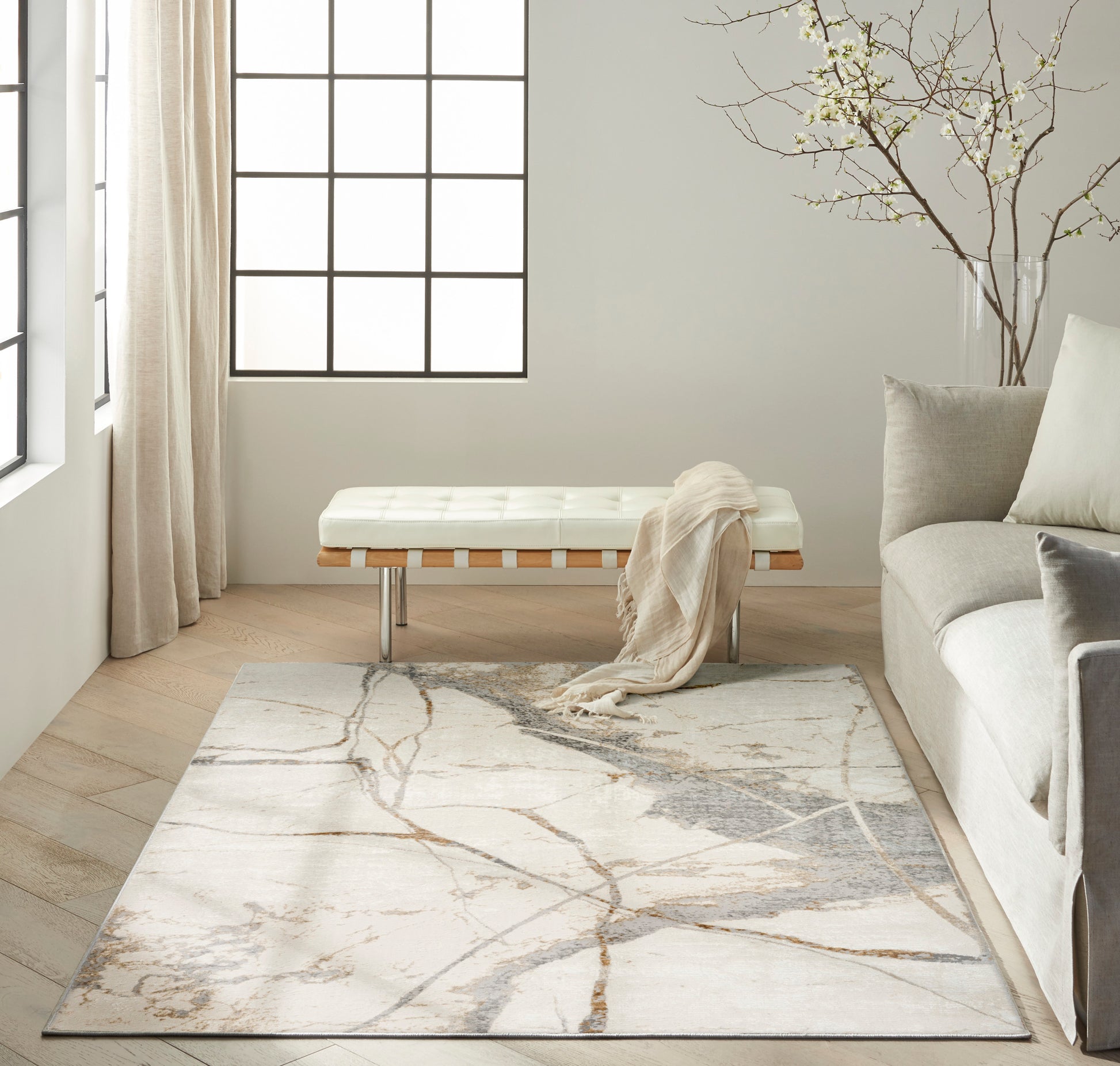 Neutral living room with white tufted bench, beige throw, gray sofa, marble-vein rug, light wood floor, large grid window.