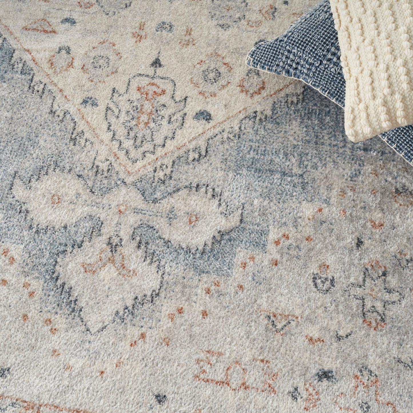Soft blue and ivory patterned area rug with subtle orange accents, paired with denim cushions and a chunky cream knit throw.