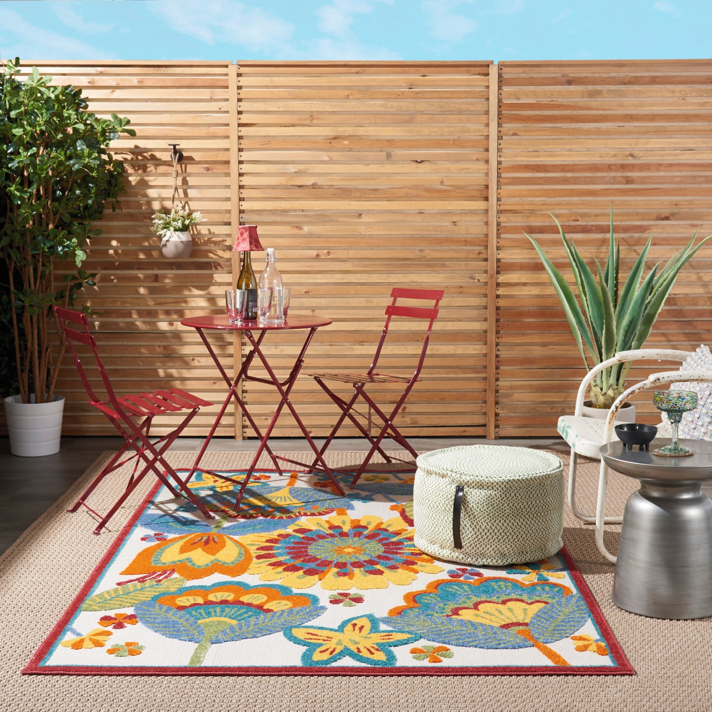 Outdoor patio with red metal bistro table and chairs, vibrant floral rug, wicker ottoman, potted plants, and wooden fence.