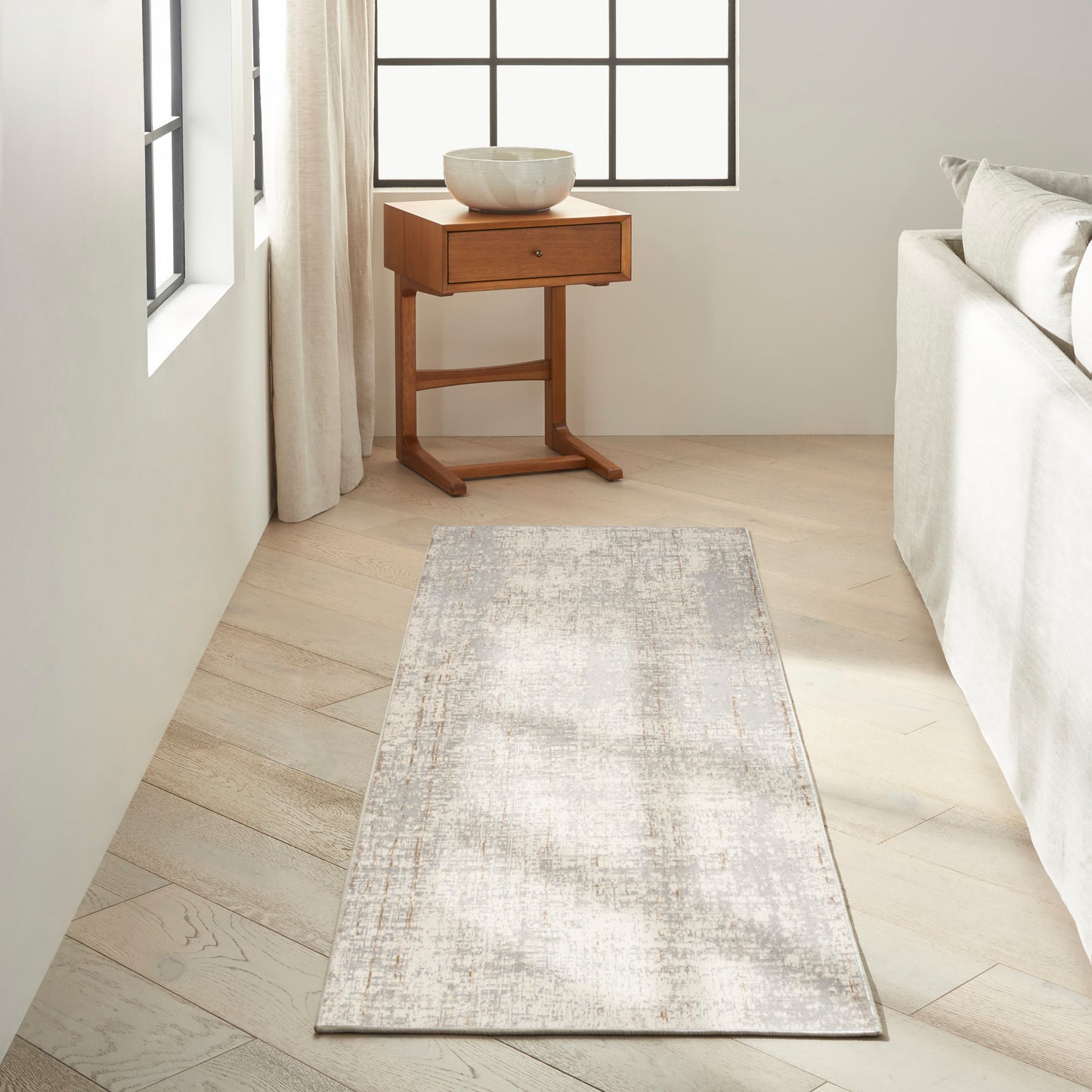 Neutral-toned living room with pale wood floors, grey distressed rug runner, round ceramic bowl on a small wooden side table near a light sofa.
