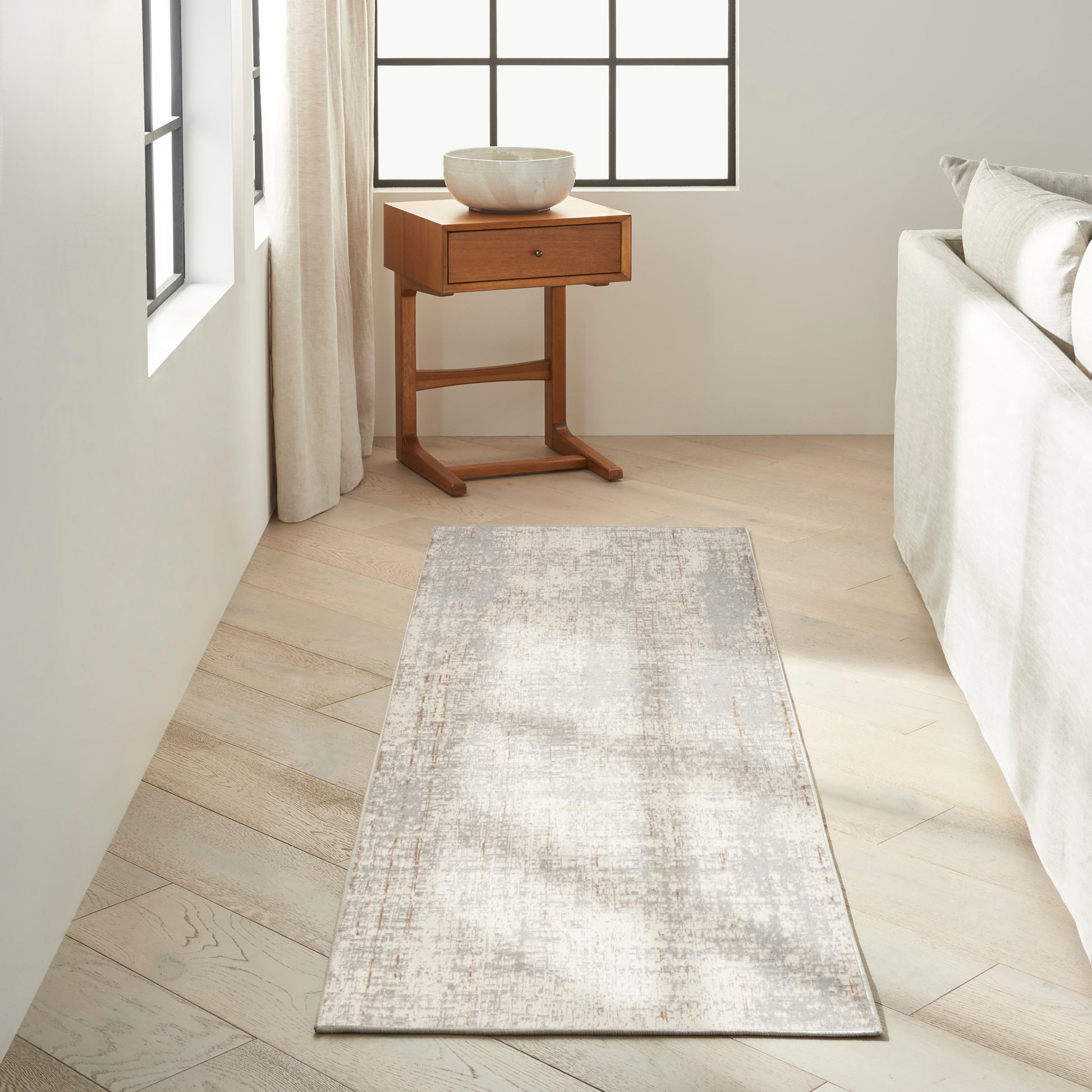 Neutral-toned living room with pale wood floors, grey distressed rug runner, round ceramic bowl on a small wooden side table near a light sofa.