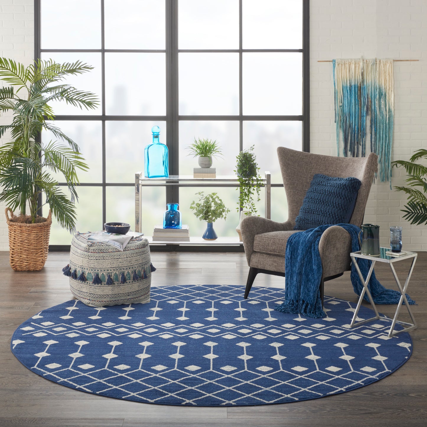Round blue geometric rug in a bright living room with a gray armchair, blue throw, glass side table, and lush plants.