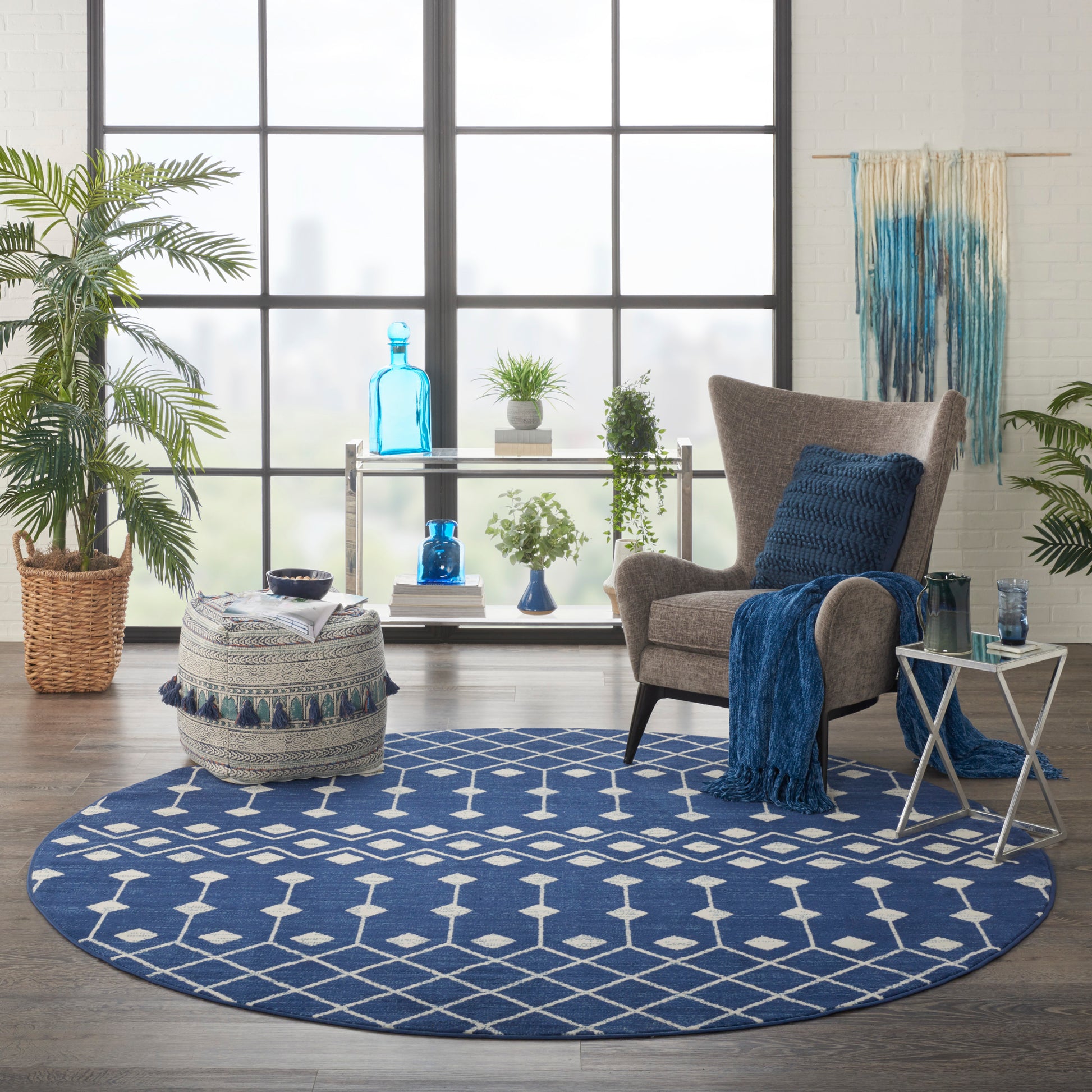 Round blue geometric rug in a bright living room with a gray armchair, blue throw, glass side table, and lush plants.