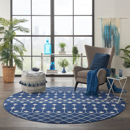 Round blue geometric rug in a bright living room with a gray armchair, blue throw, glass side table, and lush plants.