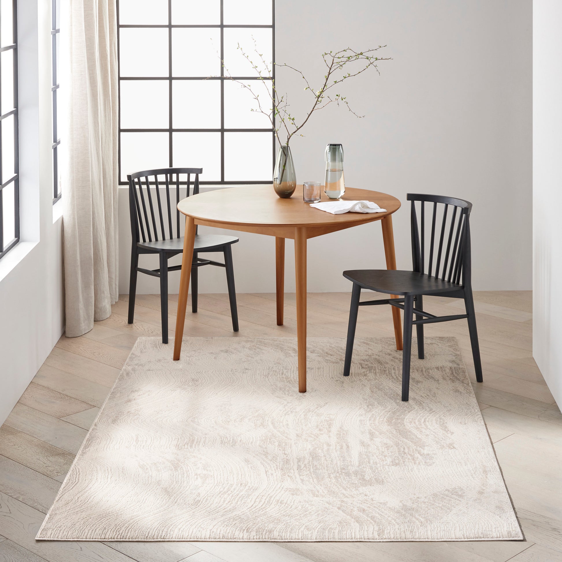 Round light-wood dining table with two black Windsor chairs, beige area rug, large grid window, vase with bare branches.
