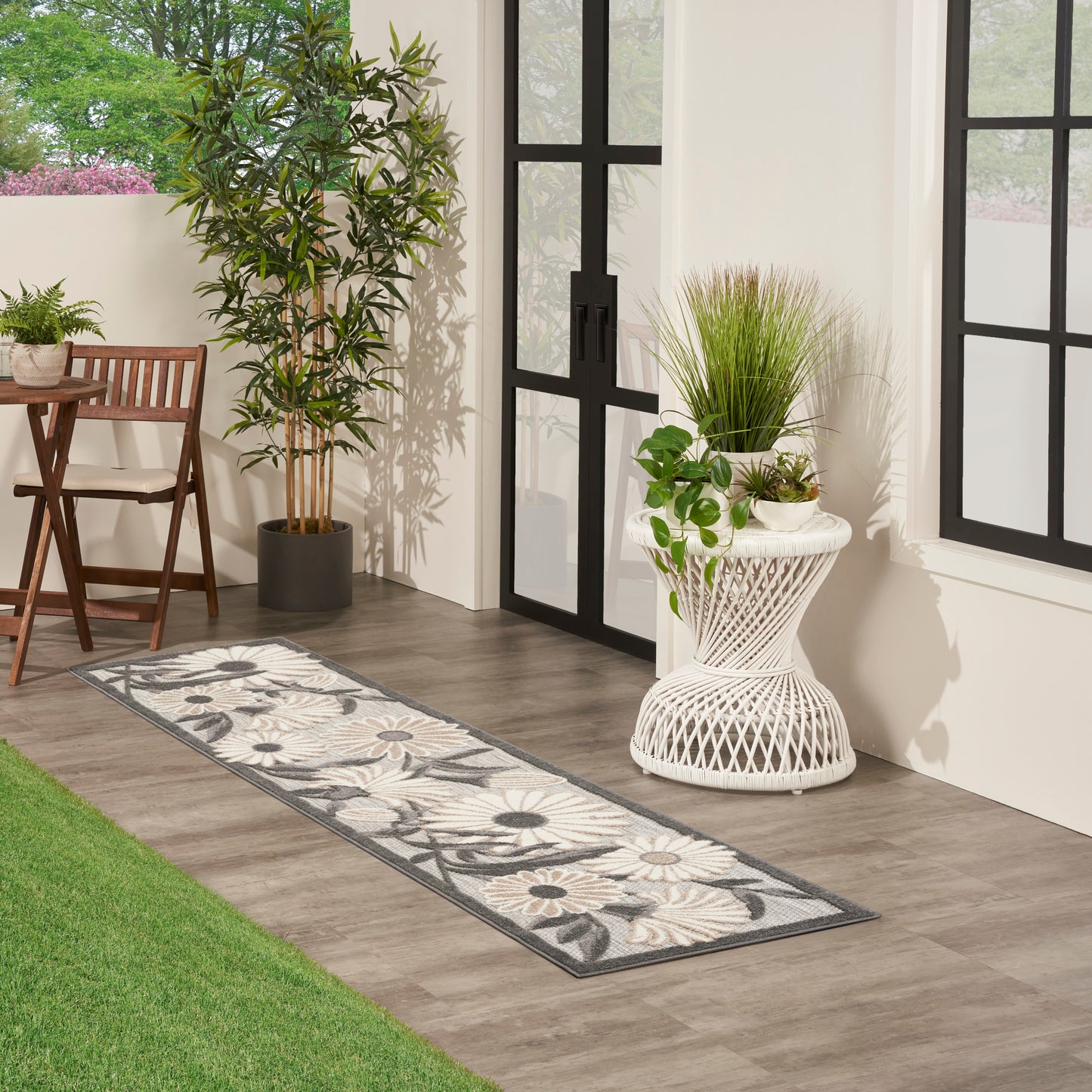 Outdoor runner rug with gray botanical pattern on wood deck beside glass doors, white wicker side table, and potted plants.