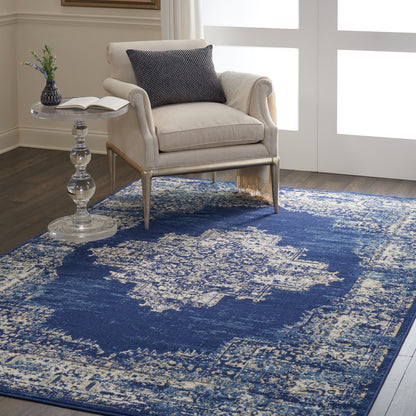 Beige upholstered armchair with tapered legs beside a glass pedestal table, navy cushion, open book, and blue distressed rug.