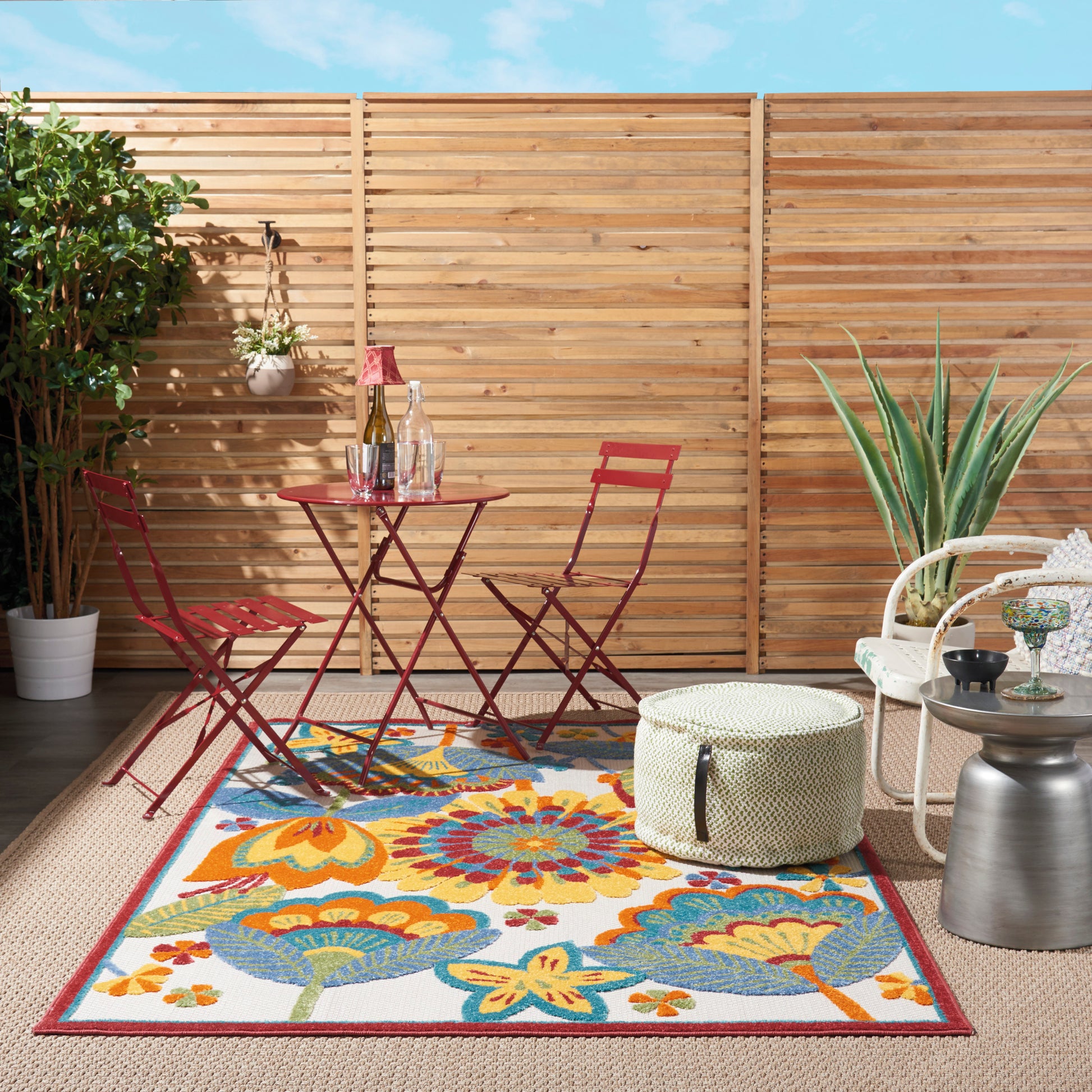 Outdoor patio with a vibrant floral rug under a red metal bistro set, wine glass on the rug; wooden fence, potted plant, wicker chair and pouf.