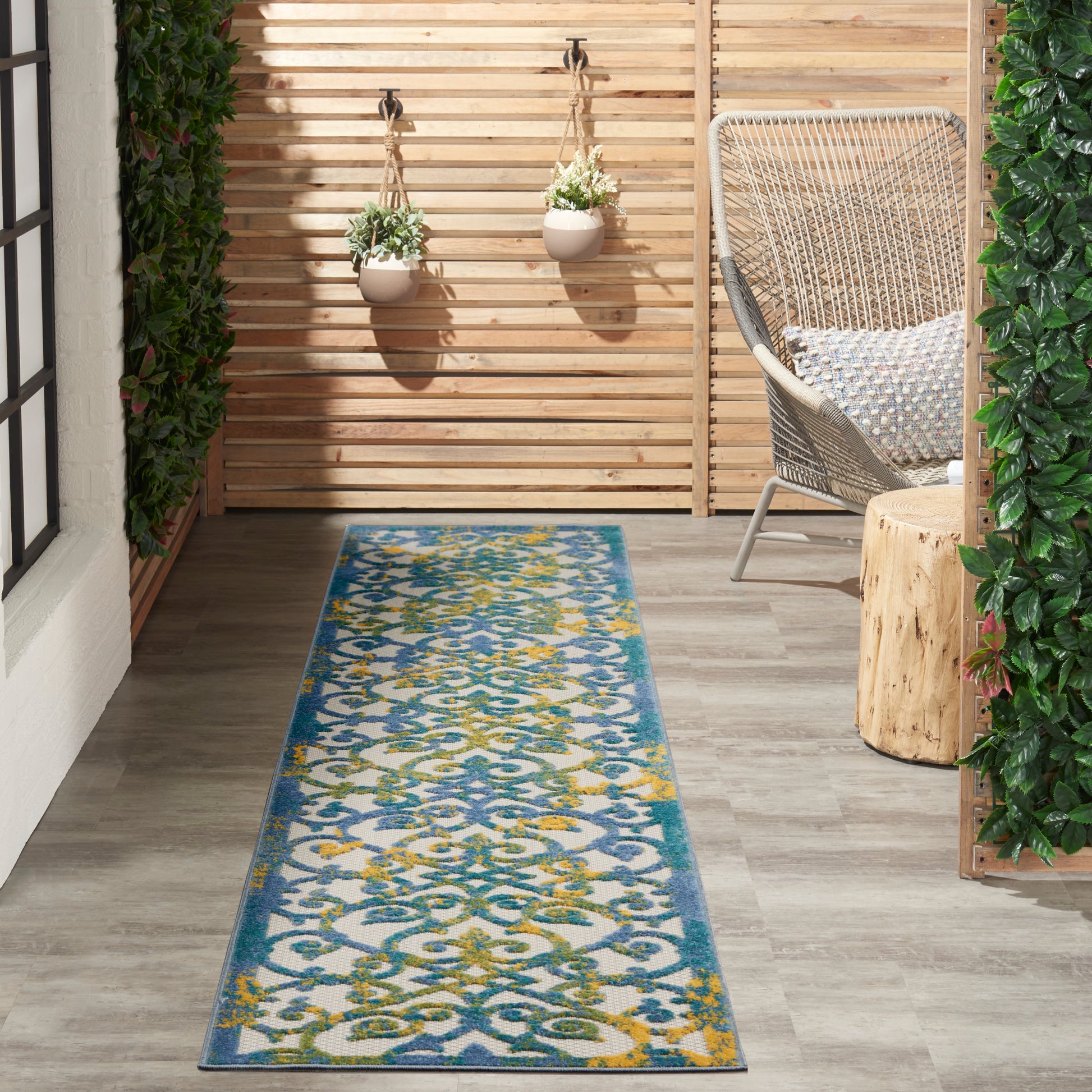 Outdoor runner rug with teal, blue, green and yellow pattern on wood deck, flanked by hanging planters, woven chair and greenery.