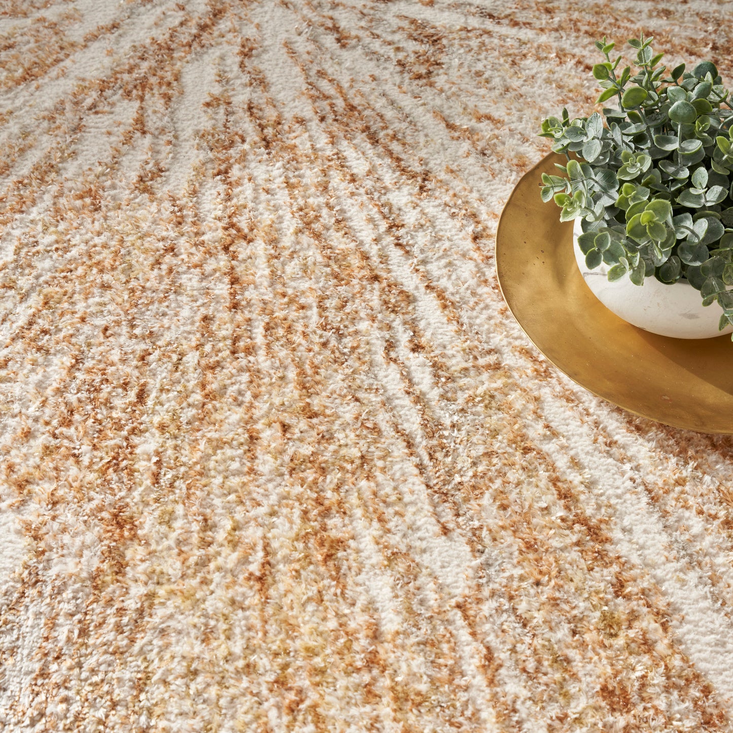 Beige cream and brown textured rug with wavy stripe pattern; round gold tray and small green plant in corner.