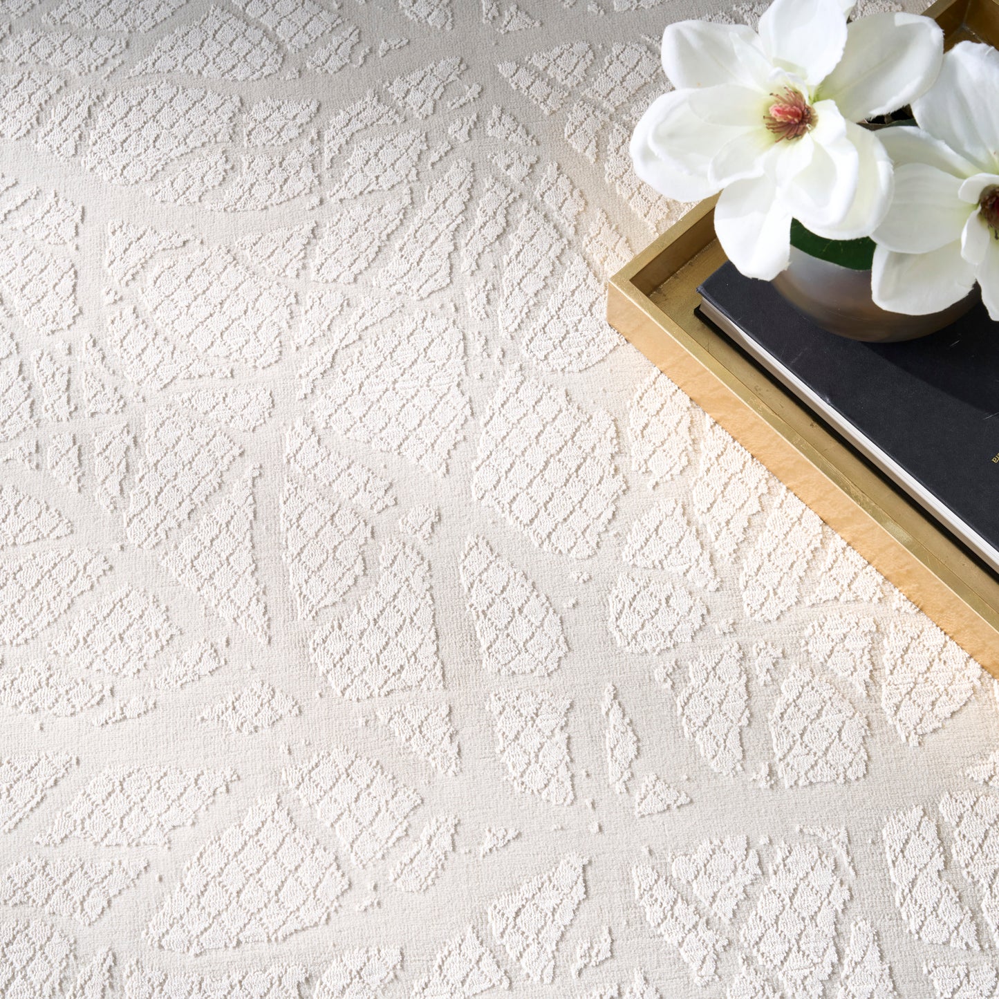 Cream textured rug with geometric leaf patterns; top-right wooden tray, black book, and white magnolia flowers.