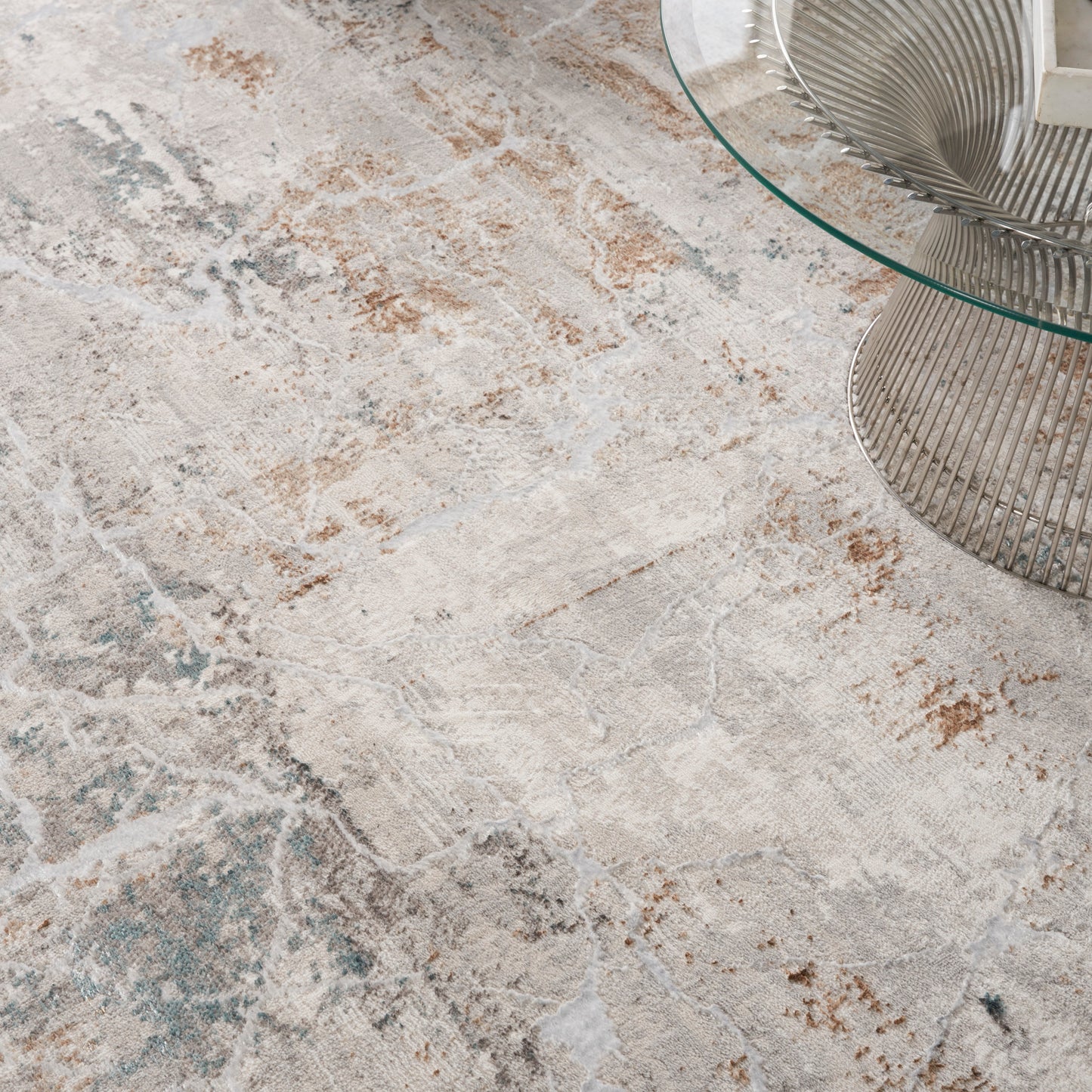 Abstract beige area rug with cream, taupe and teal accents beside a round glass coffee table with a metal wire base.