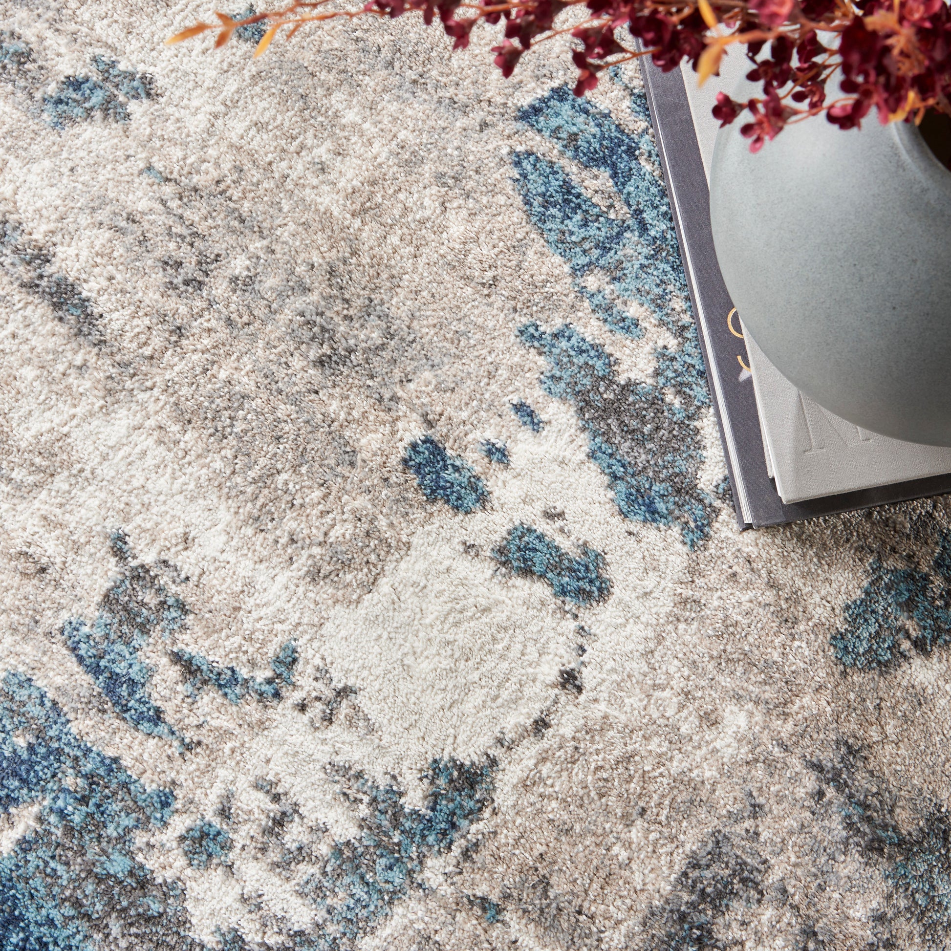 Top-down view of beige and blue abstract patterned area rug with a gray vase and red flowers atop magazines.