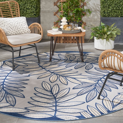 Outdoor patio with a round blue leaf-pattern rug, rattan chairs, a wooden coffee table, and potted greenery.