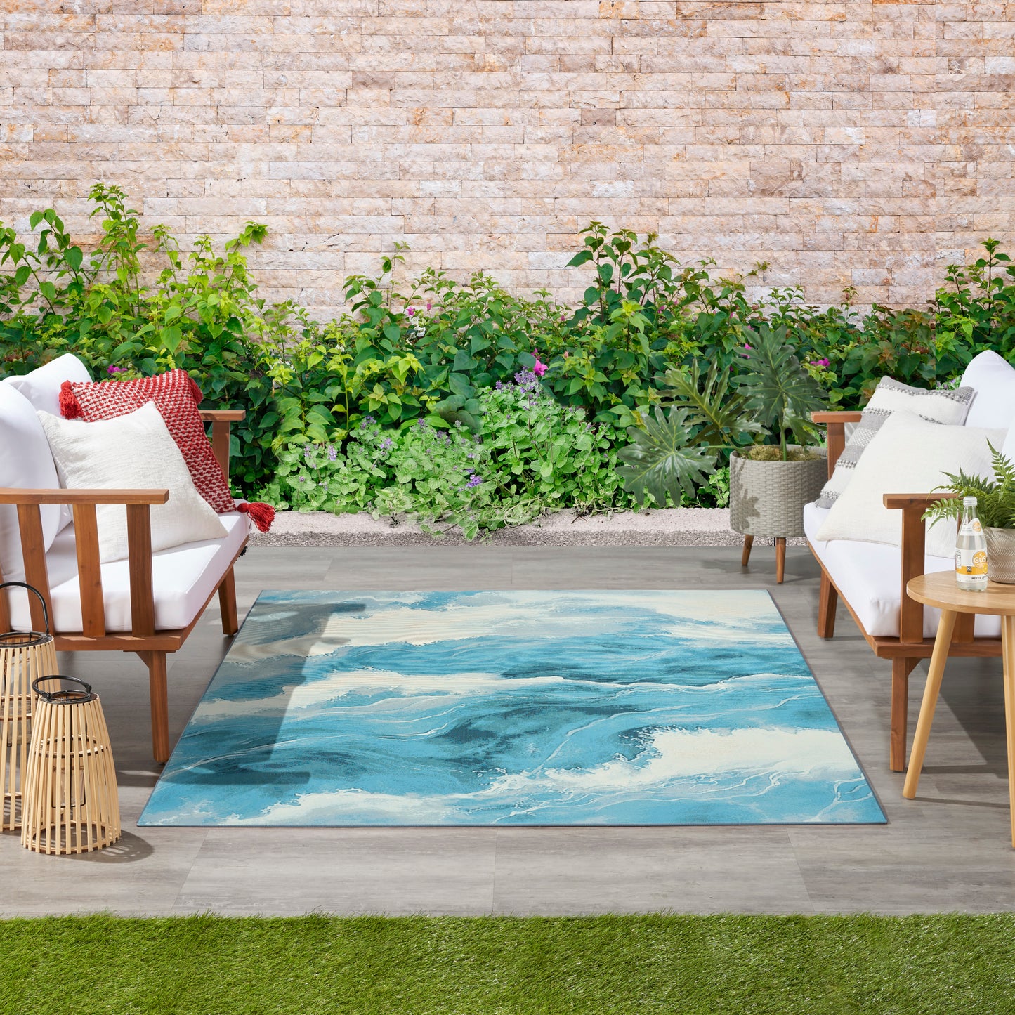 Outdoor seating area with a rectangular ocean-wave blue rug, white cushioned wooden lounge chairs, greenery and lanterns.