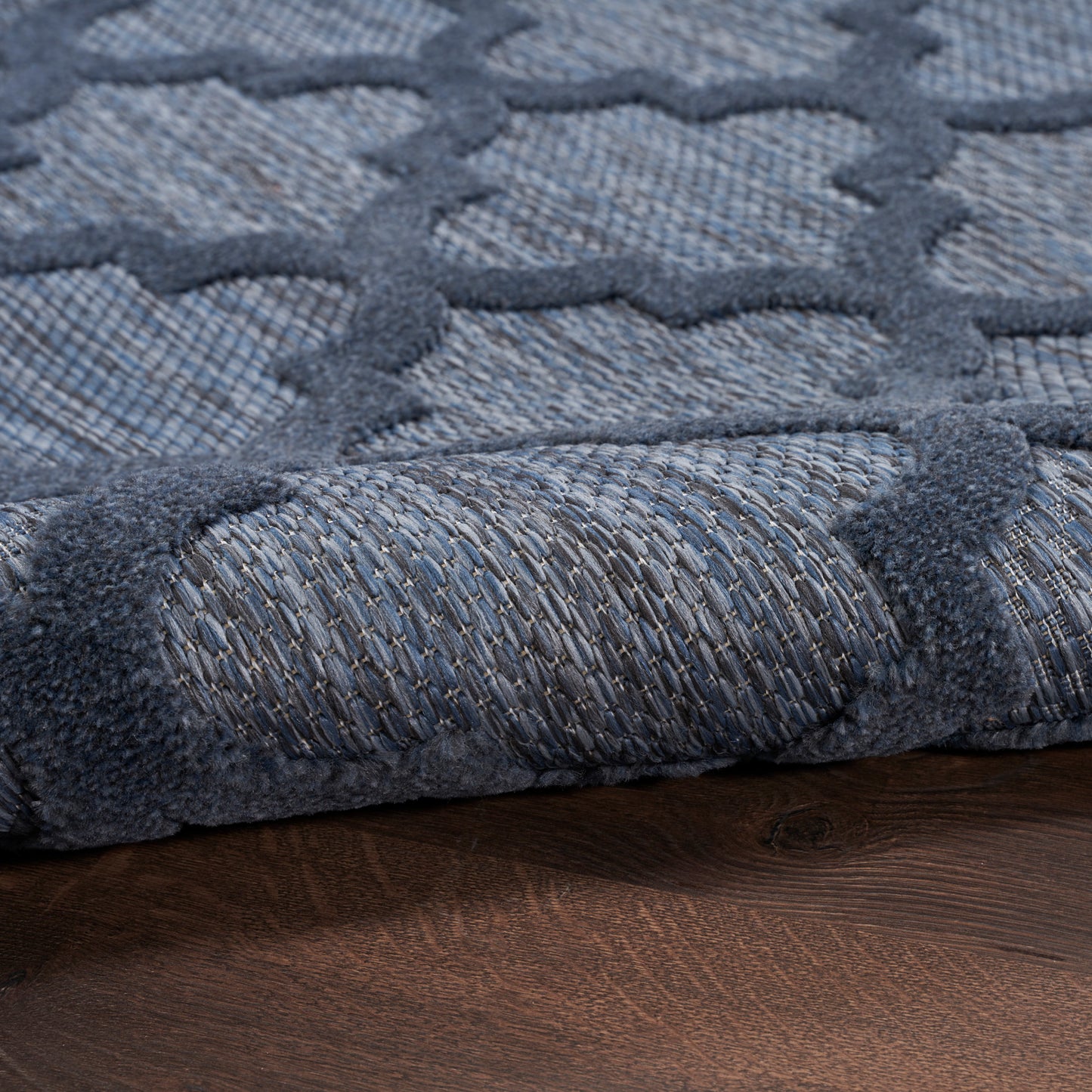 Blue textured area rug with geometric lattice pattern and rolled edge on dark wooden floor.