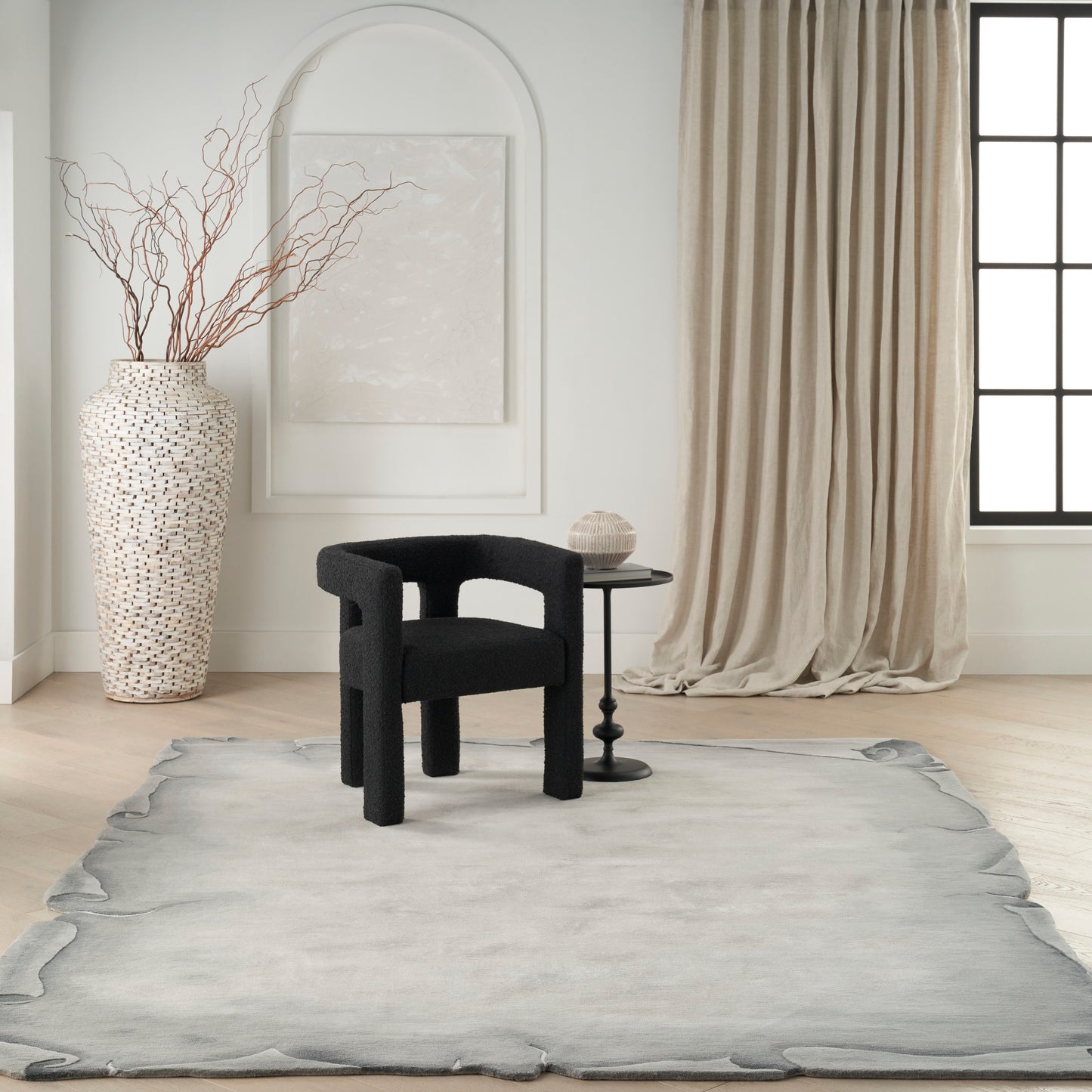 Minimalist living room with black curved-armchair, round side table, tall perforated vase with branches, and beige rug with drapery.