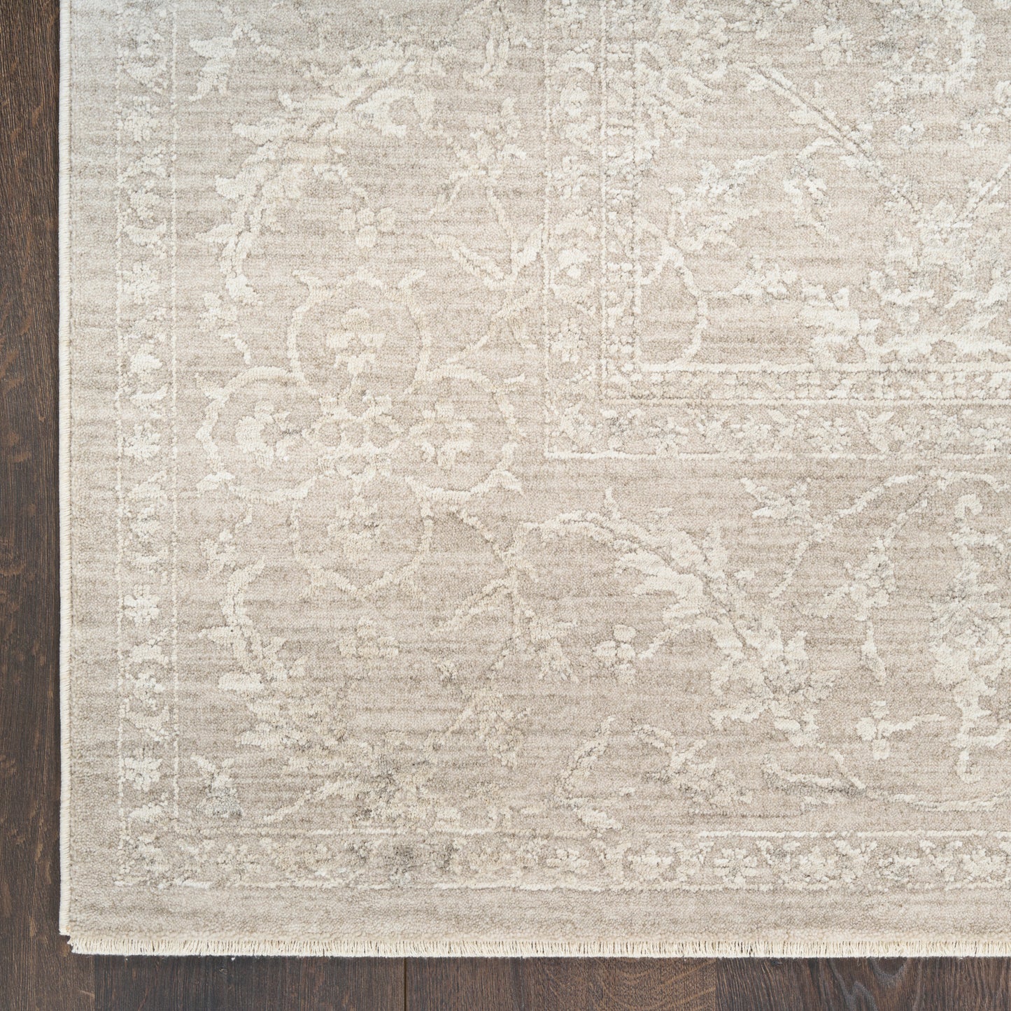 Cream-beige area rug with subtle floral pattern, ornate border and fringe on a dark wooden floor.