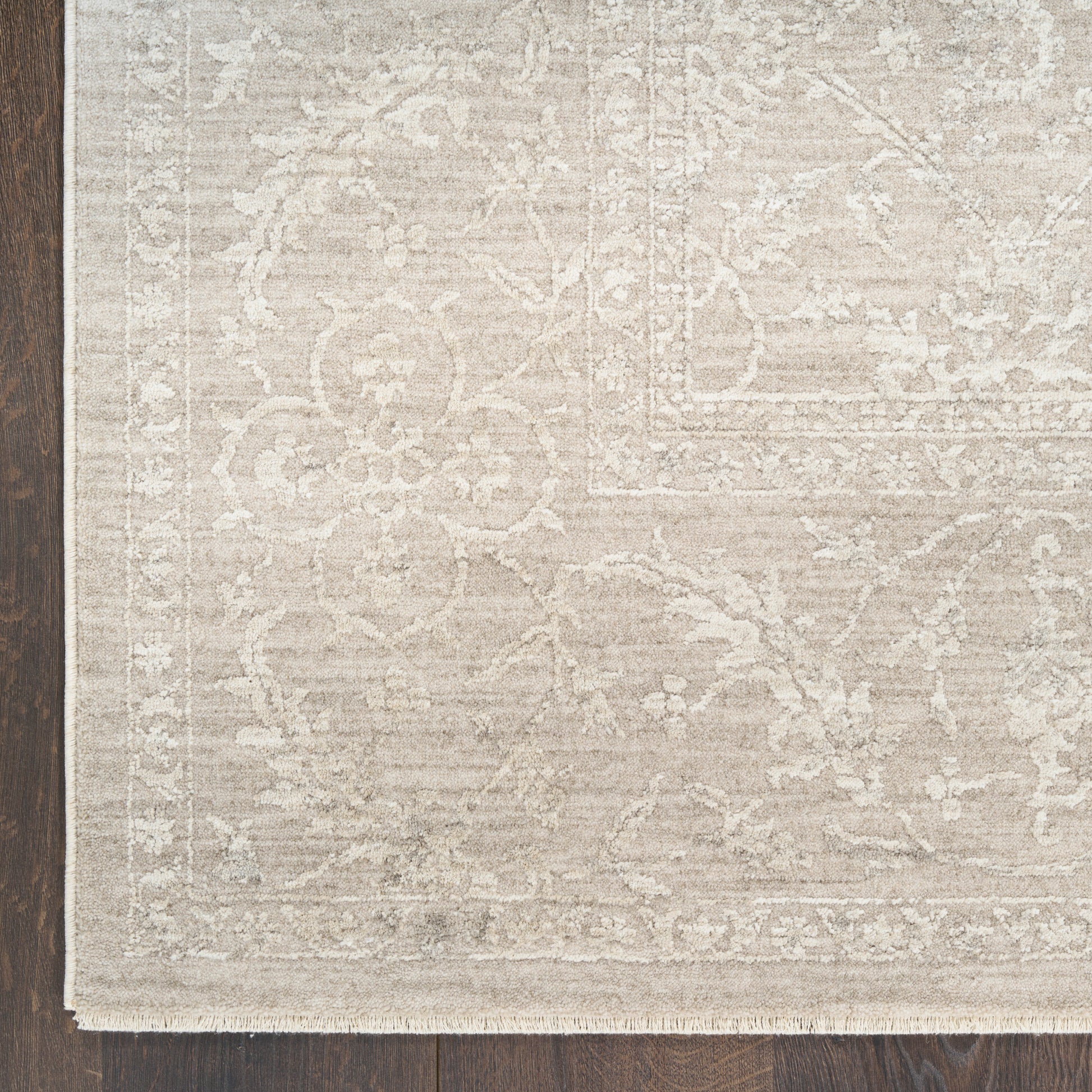 Cream-beige area rug with subtle floral pattern, ornate border and fringe on a dark wooden floor.