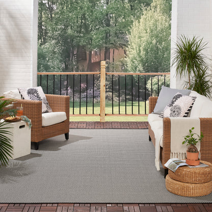 Gray rug on a brick porch under outdoor wicker seating with white cushions, patterned pillows, a round ottoman, potted plants, and a garden view.