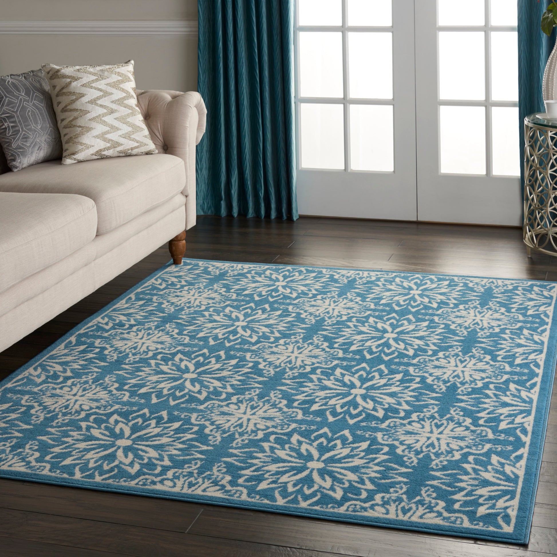 Blue damask-patterned area rug with white floral motifs and a teal border, in a living room with beige sofa and teal curtains.