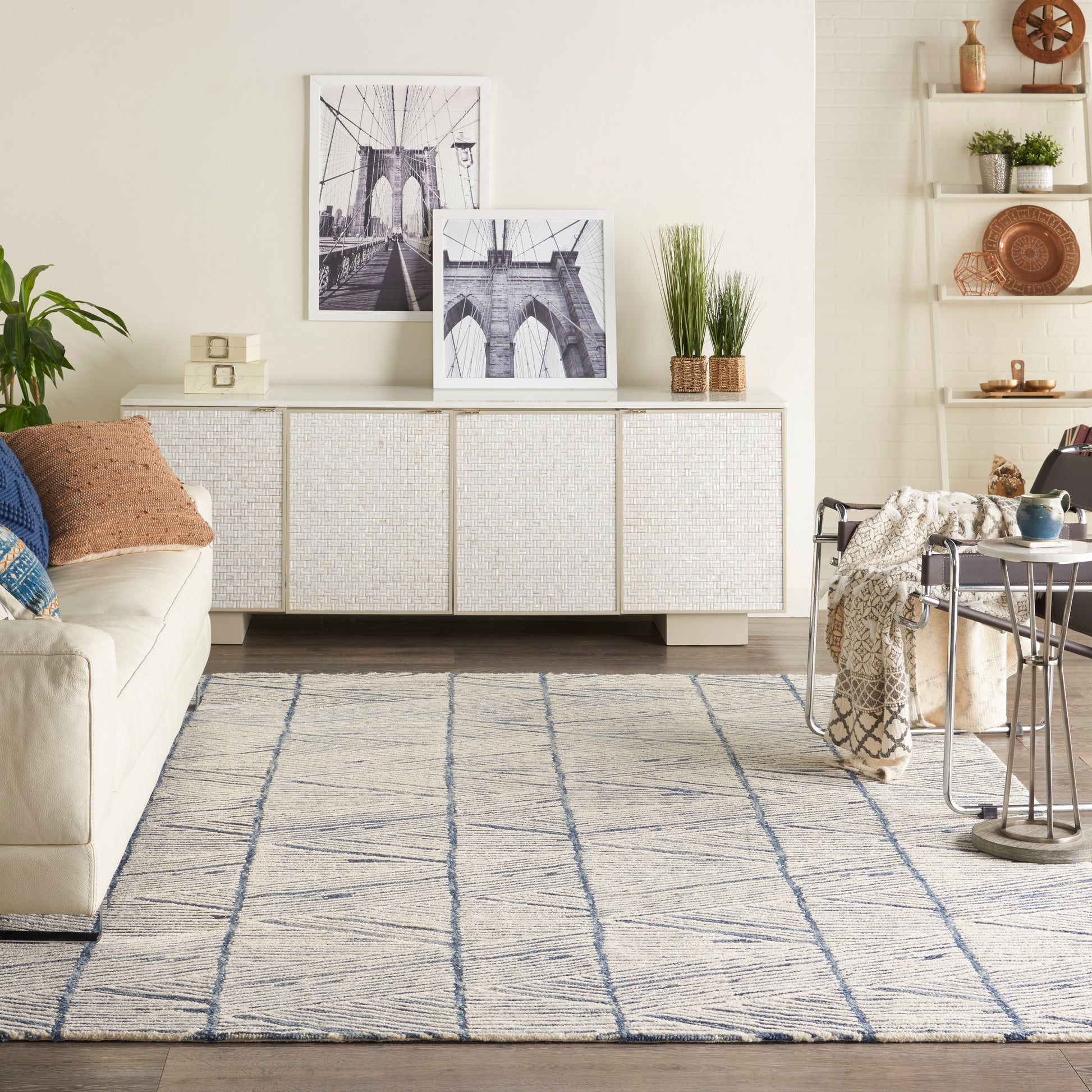 Neutral living room with cream sofa, woven white cabinet, black-and-white bridge photos, blue patterned rug, plants.