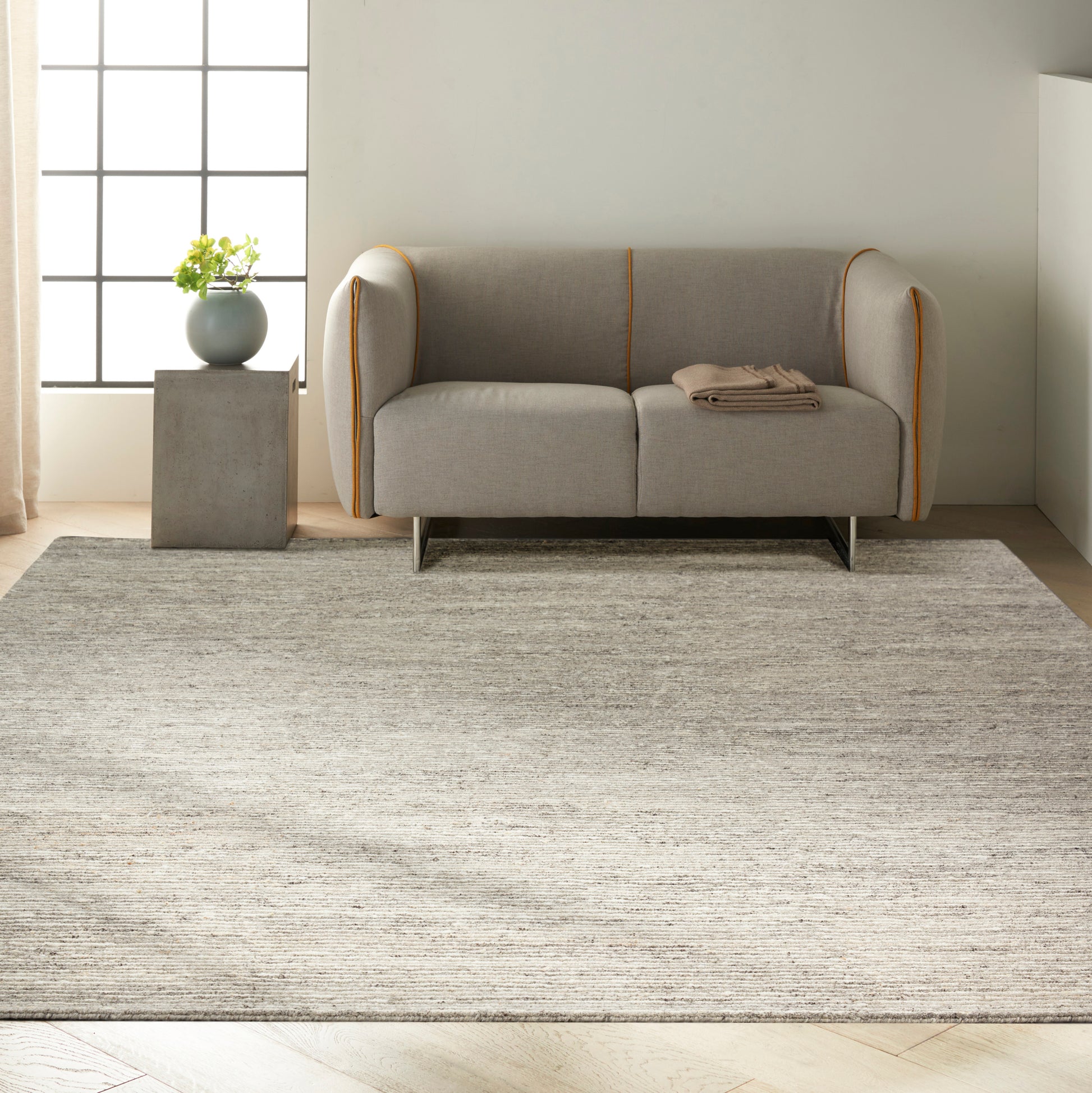 Gray textured rug; a gray fabric sofa with orange piping and rounded arms sits on it, beside a concrete side table with a potted plant.