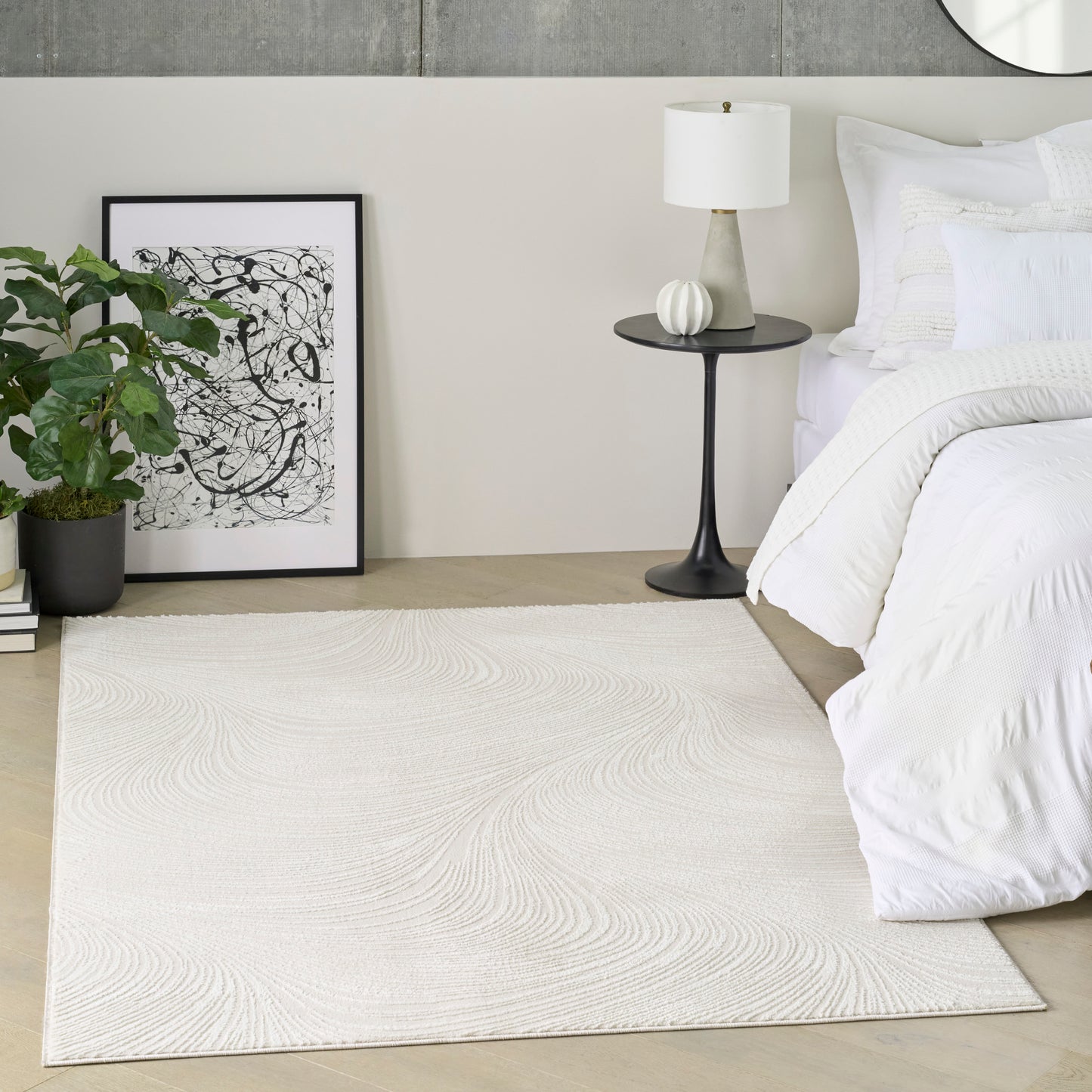 Neutral bedroom with white duvet, black nightstand and lamp, beige textured area rug, potted plant, and framed abstract art.