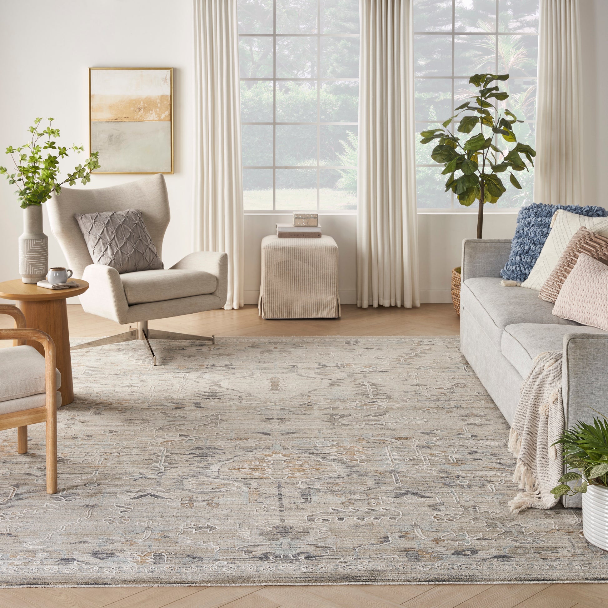 Neutral living room with beige fabric armchair, gray sofa, patterned rug, greenery, and large window with cream curtains.