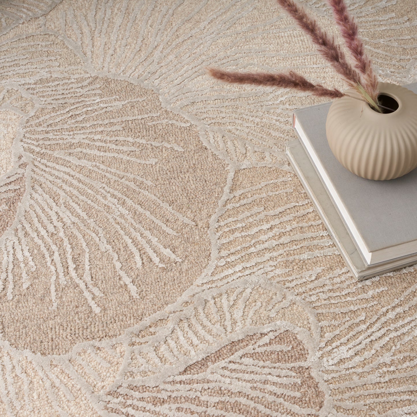 Beige textured rug with large leaf motif, paired with a ribbed cream vase, pampas grass, and stacked books.
