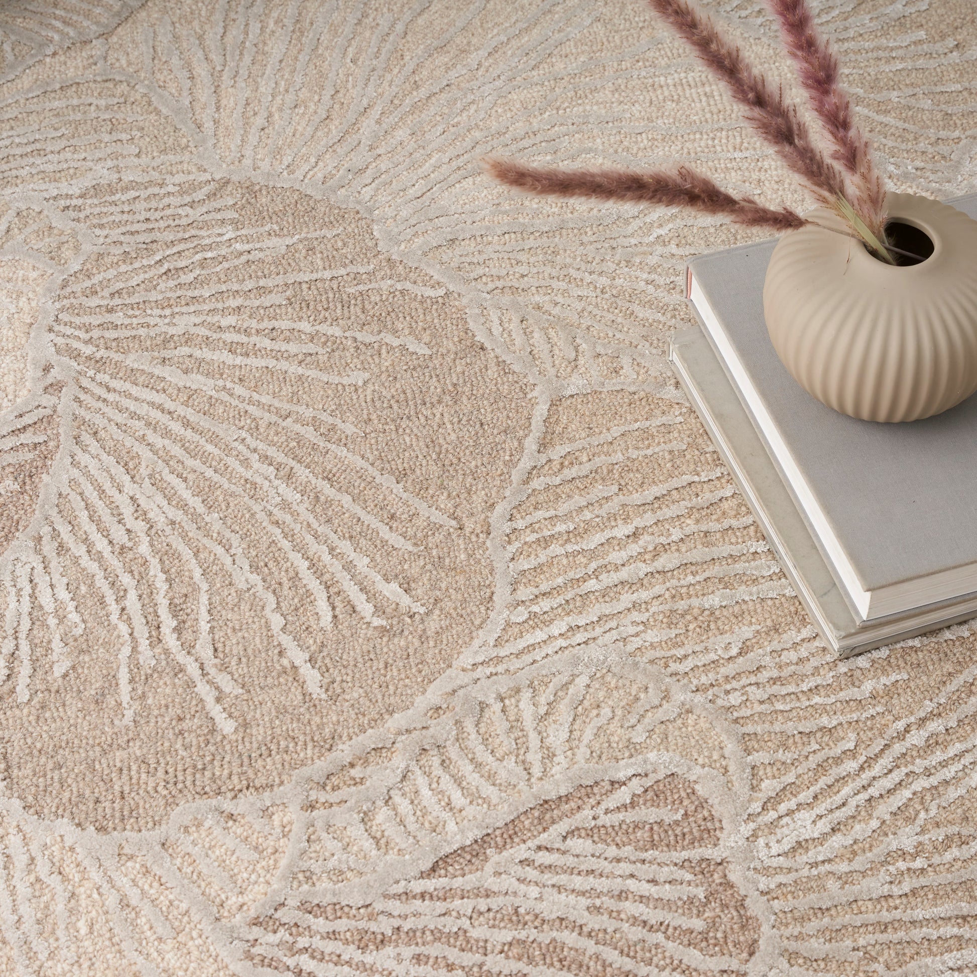 Beige textured rug with large leaf motif, paired with a ribbed cream vase, pampas grass, and stacked books.