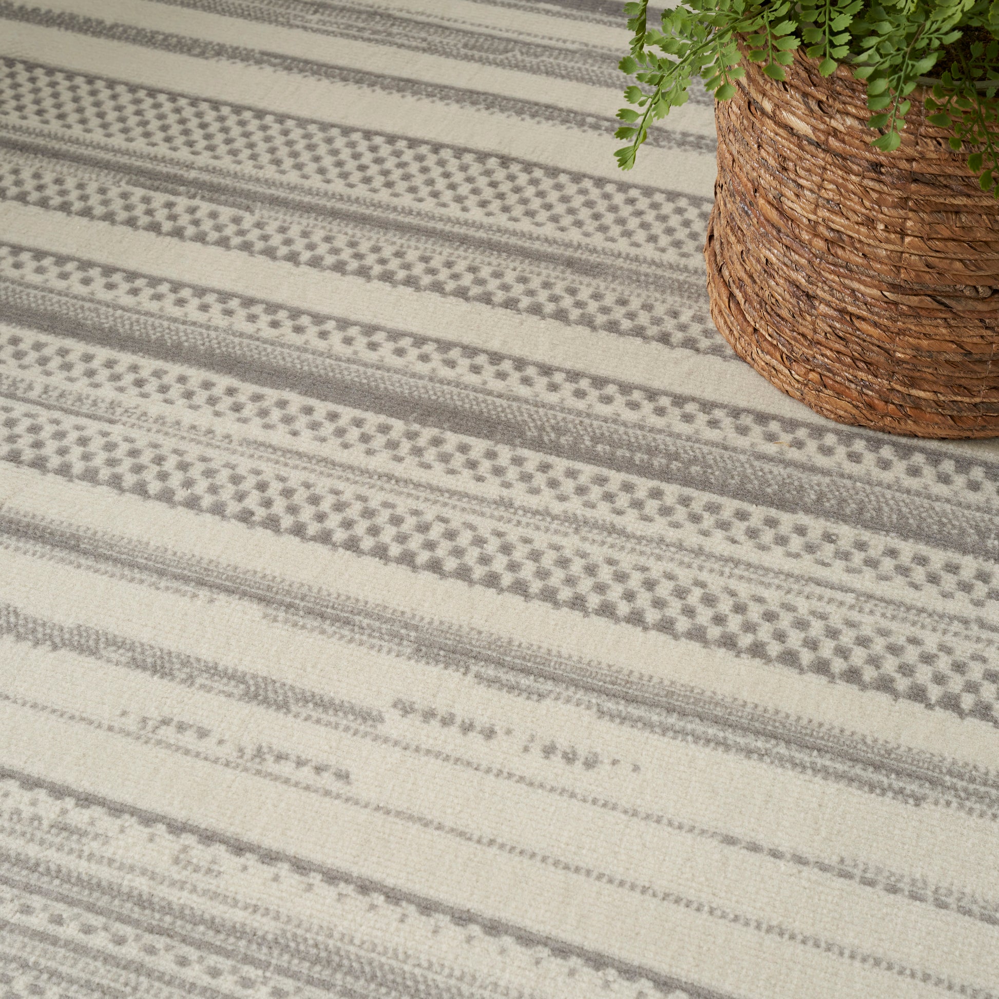 Beige & gray striped woven rug with geometric motifs, placed on the floor beside a woven jute basket with a green plant.