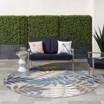 Round beige outdoor rug with blue and gray leaf pattern under navy cushioned patio sofa, side table, hedge wall backdrop.