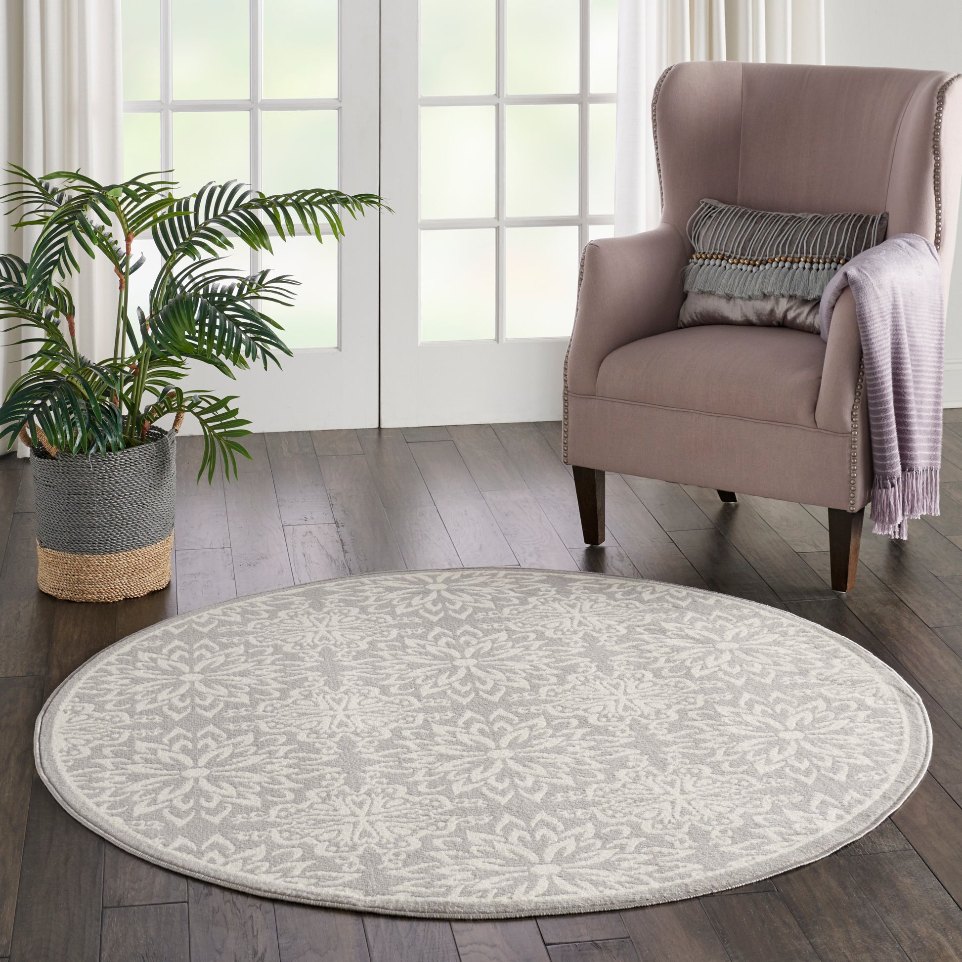 Round beige patterned area rug on dark wood floor, with pink upholstered wing chair and plant in woven basket.