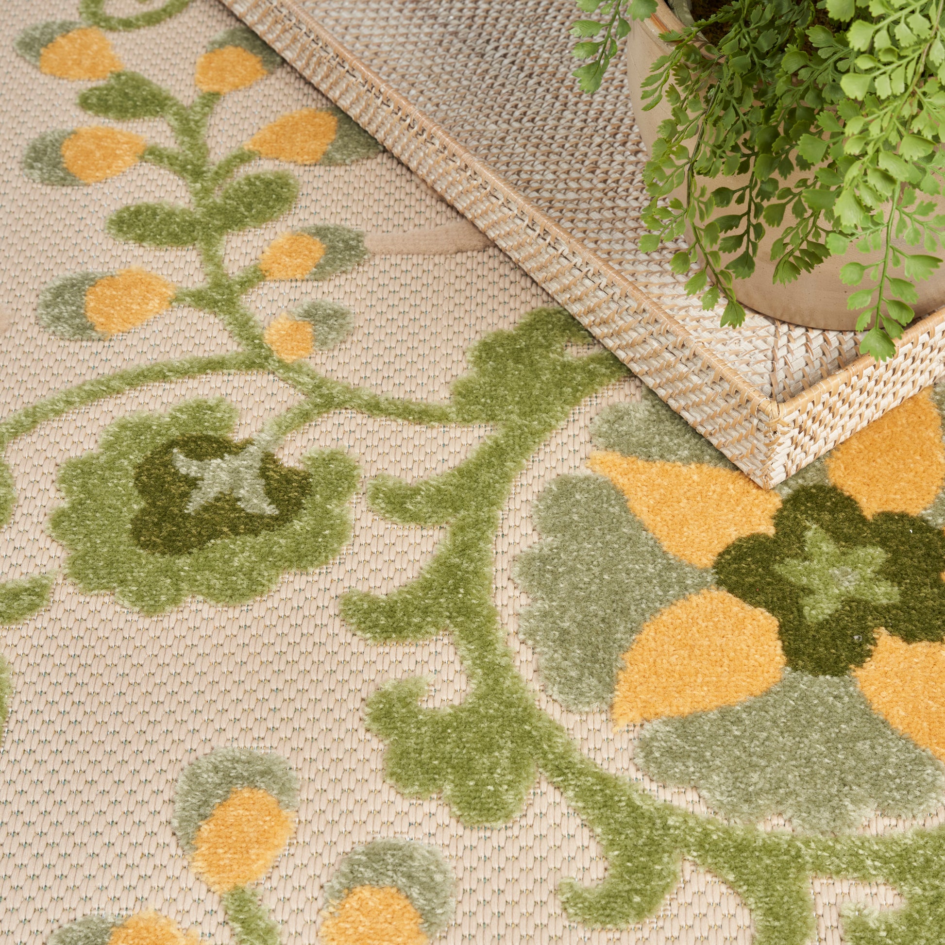 Beige woven rug with green and mustard-yellow floral motif; rattan tray and leafy plant nearby.