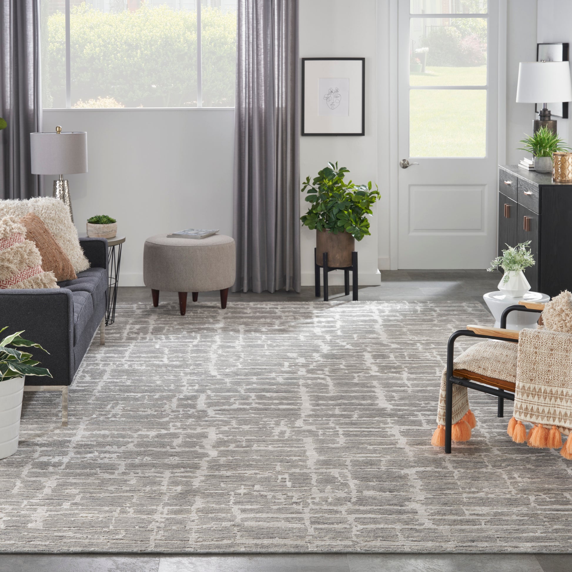 Contemporary living room with gray sofa, beige pillows, round tufted ottoman, abstract geometric rug, and lush plants.