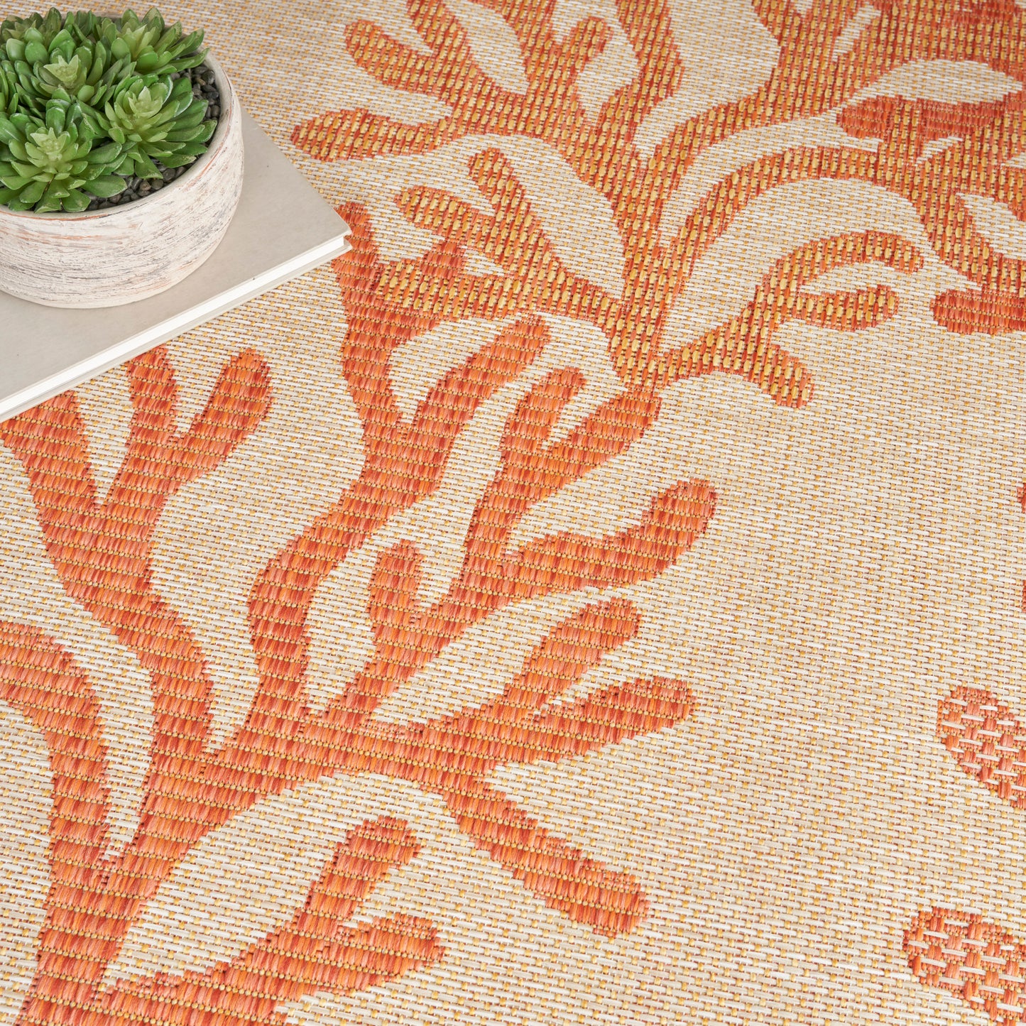 Orange coral-patterned woven rug on a beige background, with a small potted succulent atop a light book.