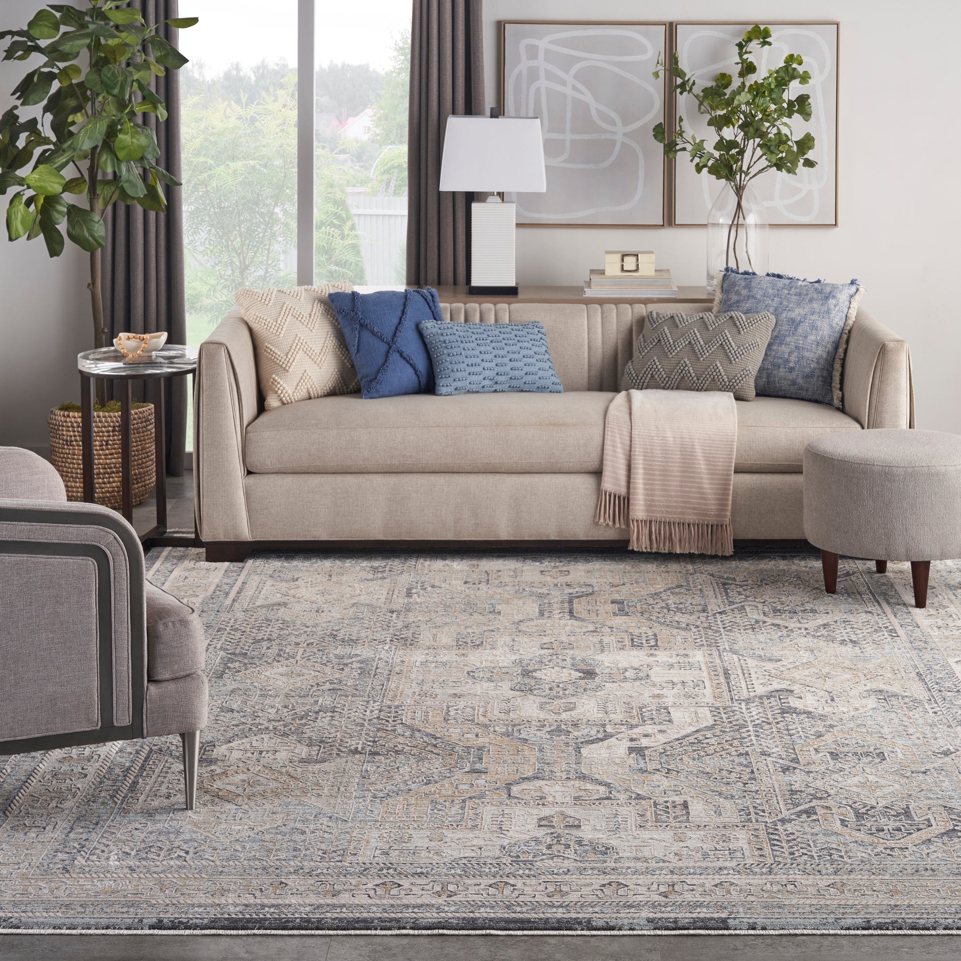 Beige fabric sofa with blue and gray textured cushions, a throw blanket, abstract wall art, plants, and a patterned rug.