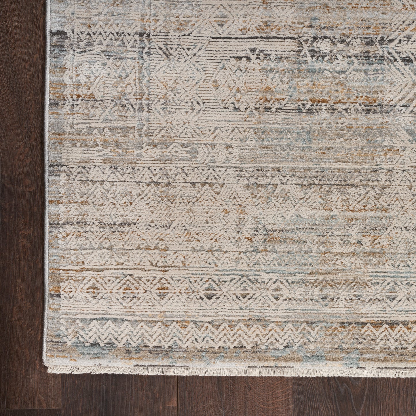Distressed geometric patterned area rug in beige, ivory, tan and muted blue with fringe along bottom edge on dark wood floor.