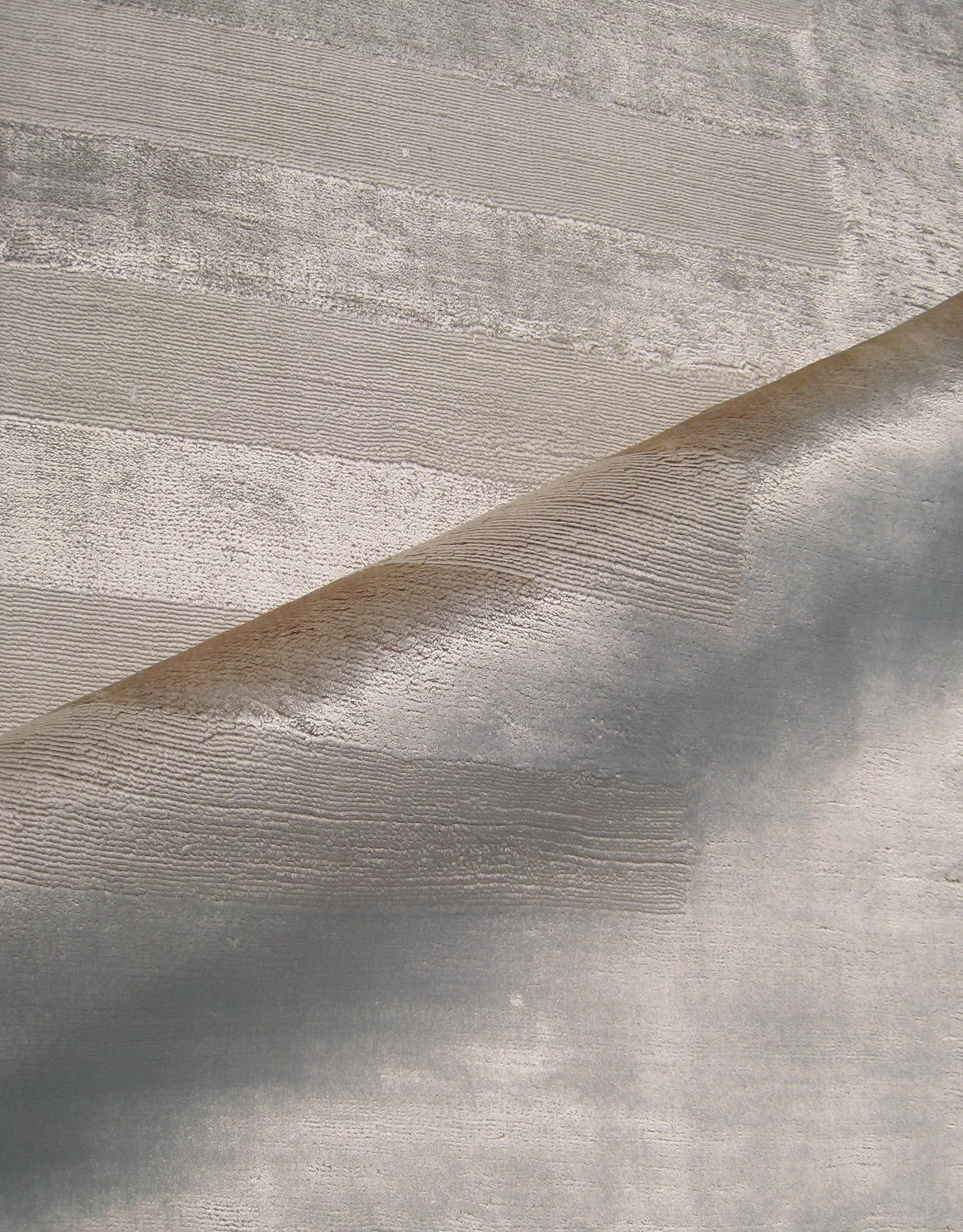 Curl of metallic beige rug with brushed horizontal stripes against a light gray textured surface.