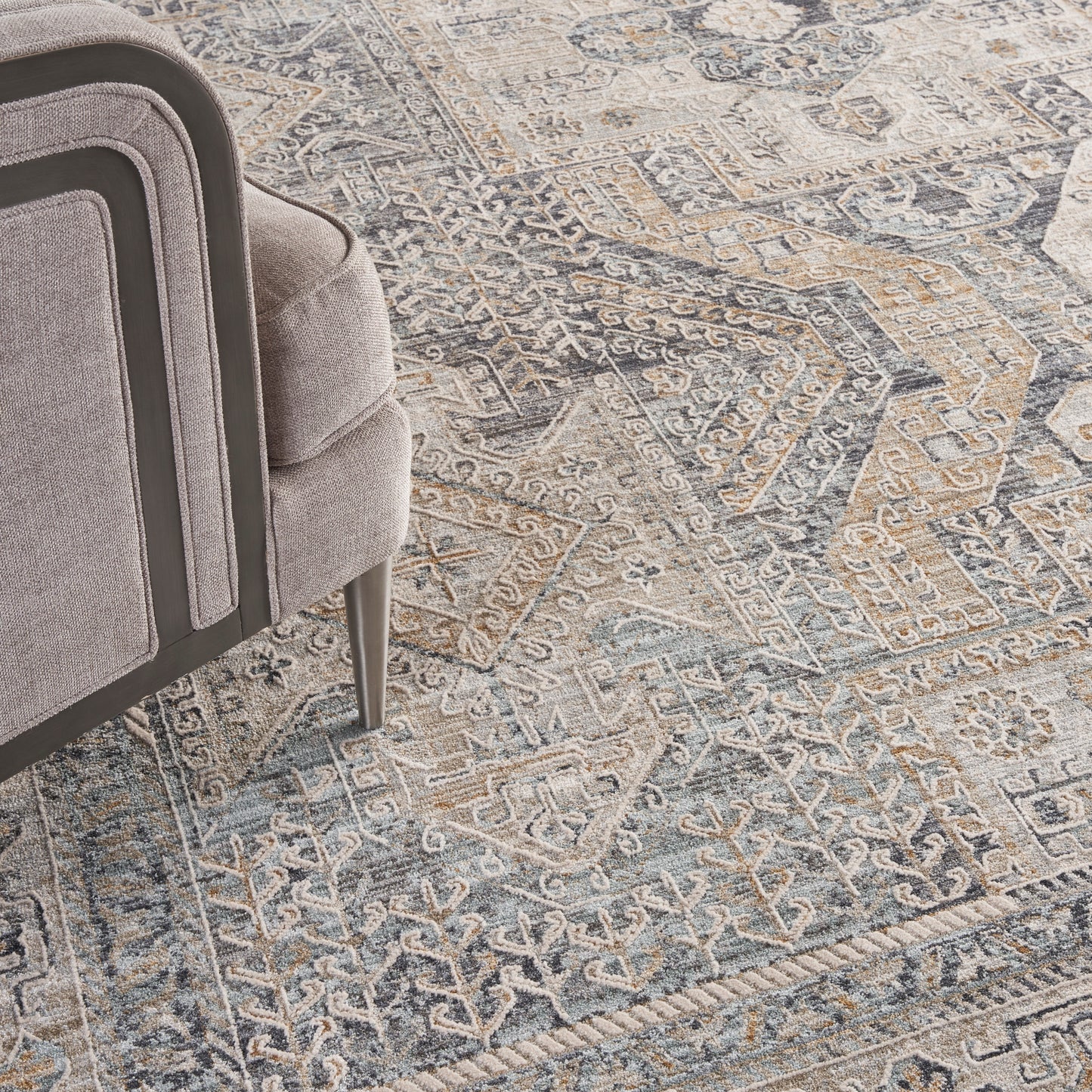Partial view of a light gray upholstered armchair with dark piping on a blue-gray geometric oriental rug